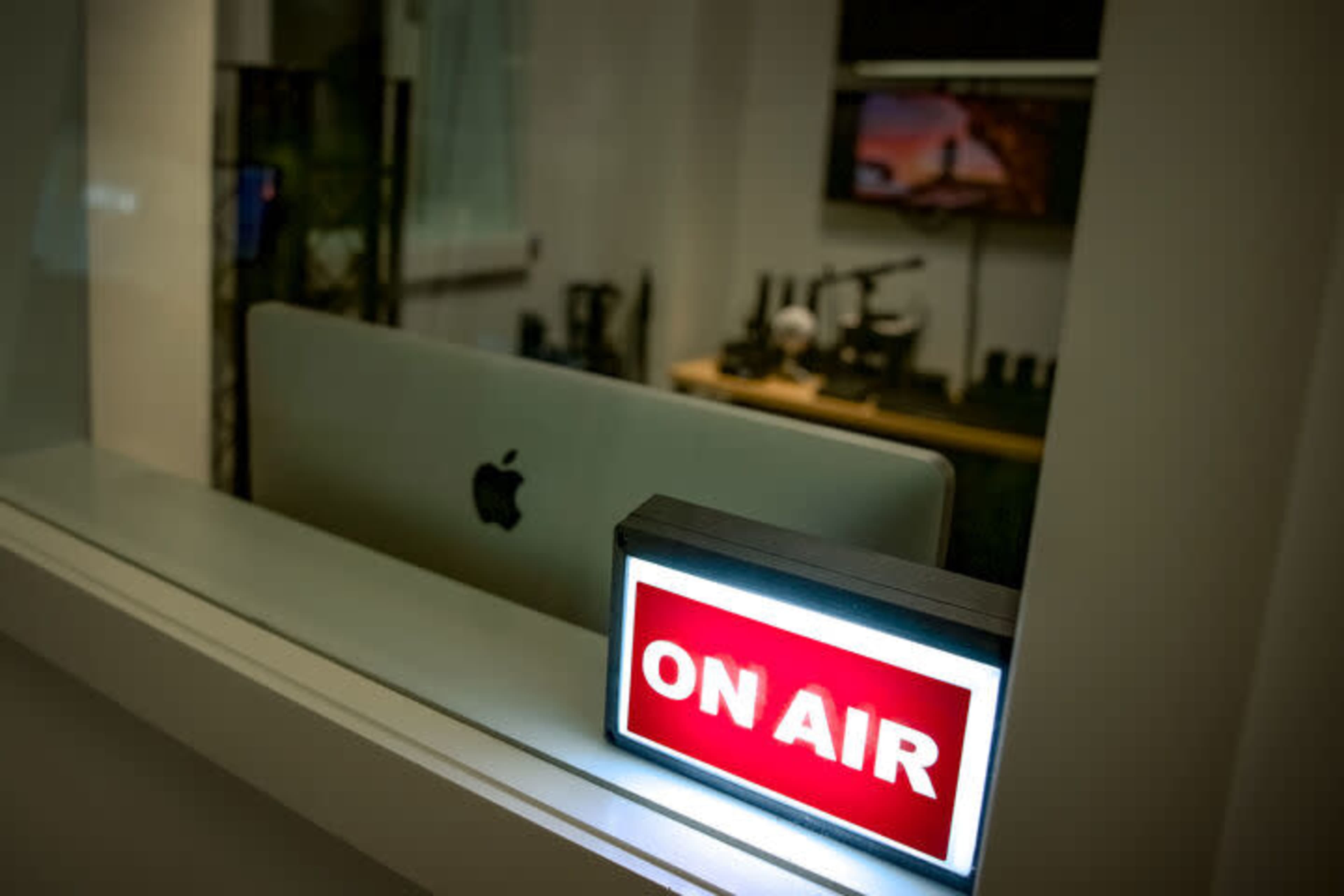 A illuminated "ON AIR" sign is displayed on a shelf near an Apple computer in a studio environment.