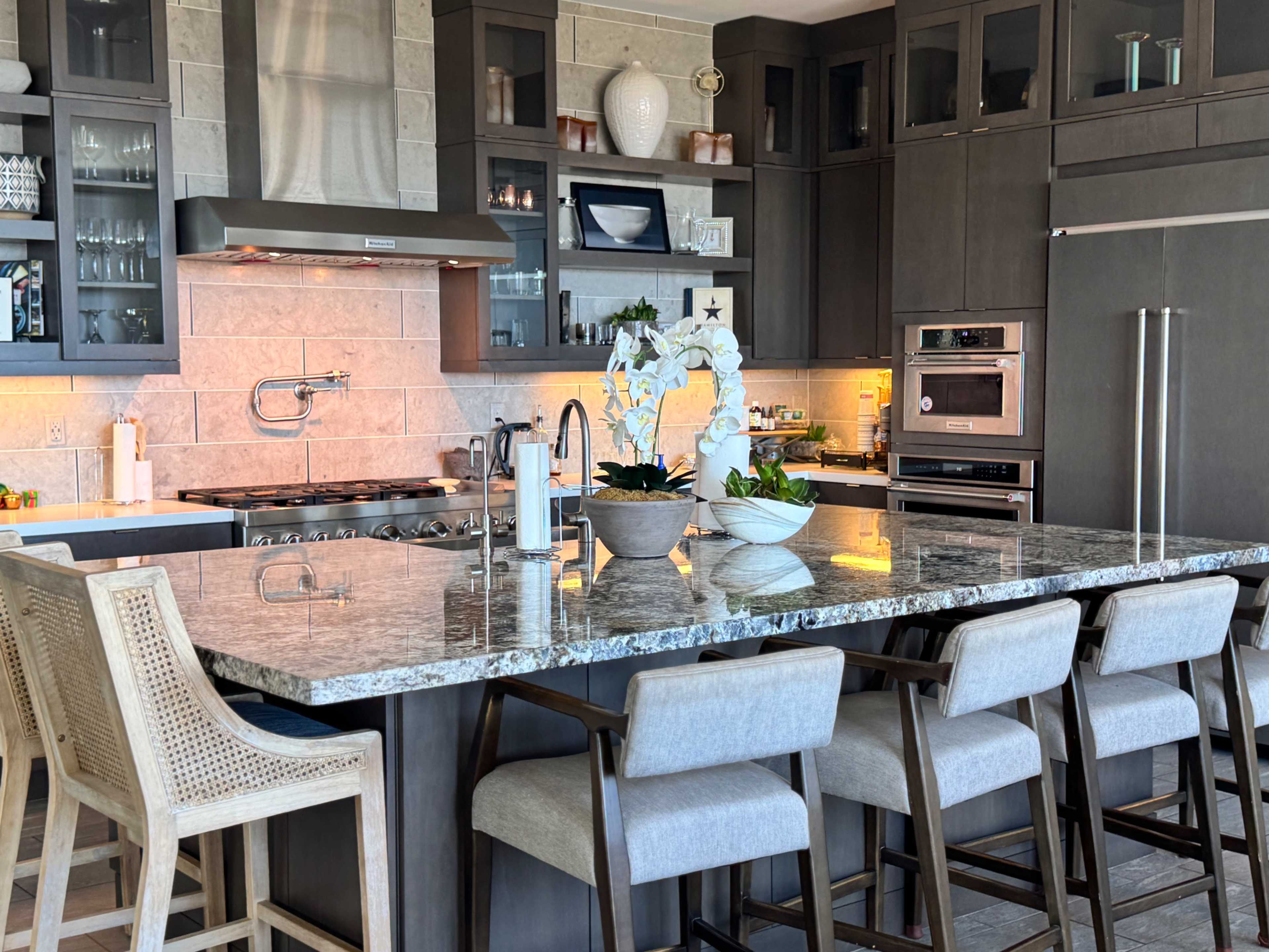 The image shows a modern kitchen with a large granite island, high-backed wooden stools, and stainless steel appliances.