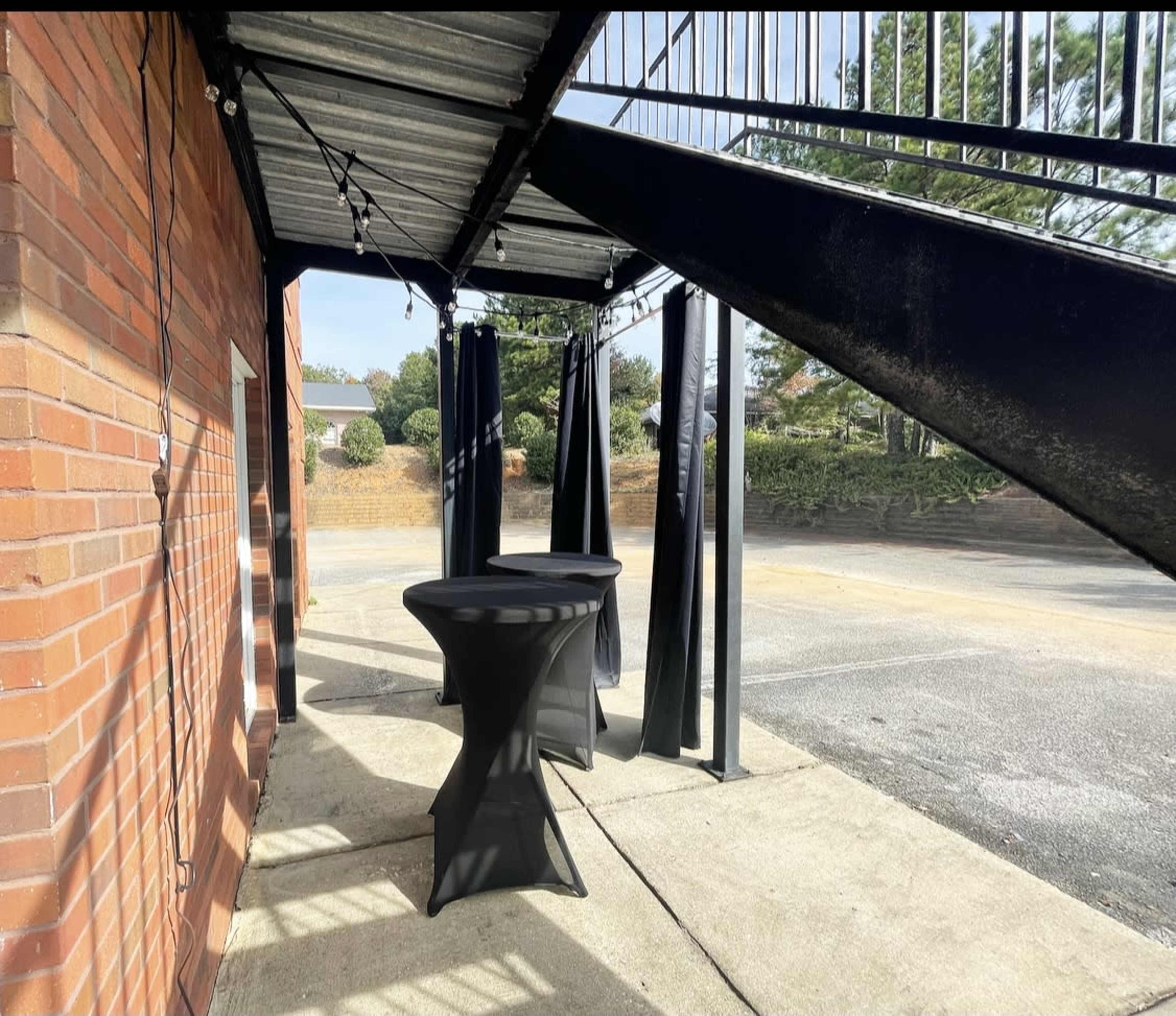 A covered outdoor space features two black tables and curtains beneath a staircase, with a parking lot visible in the background.
