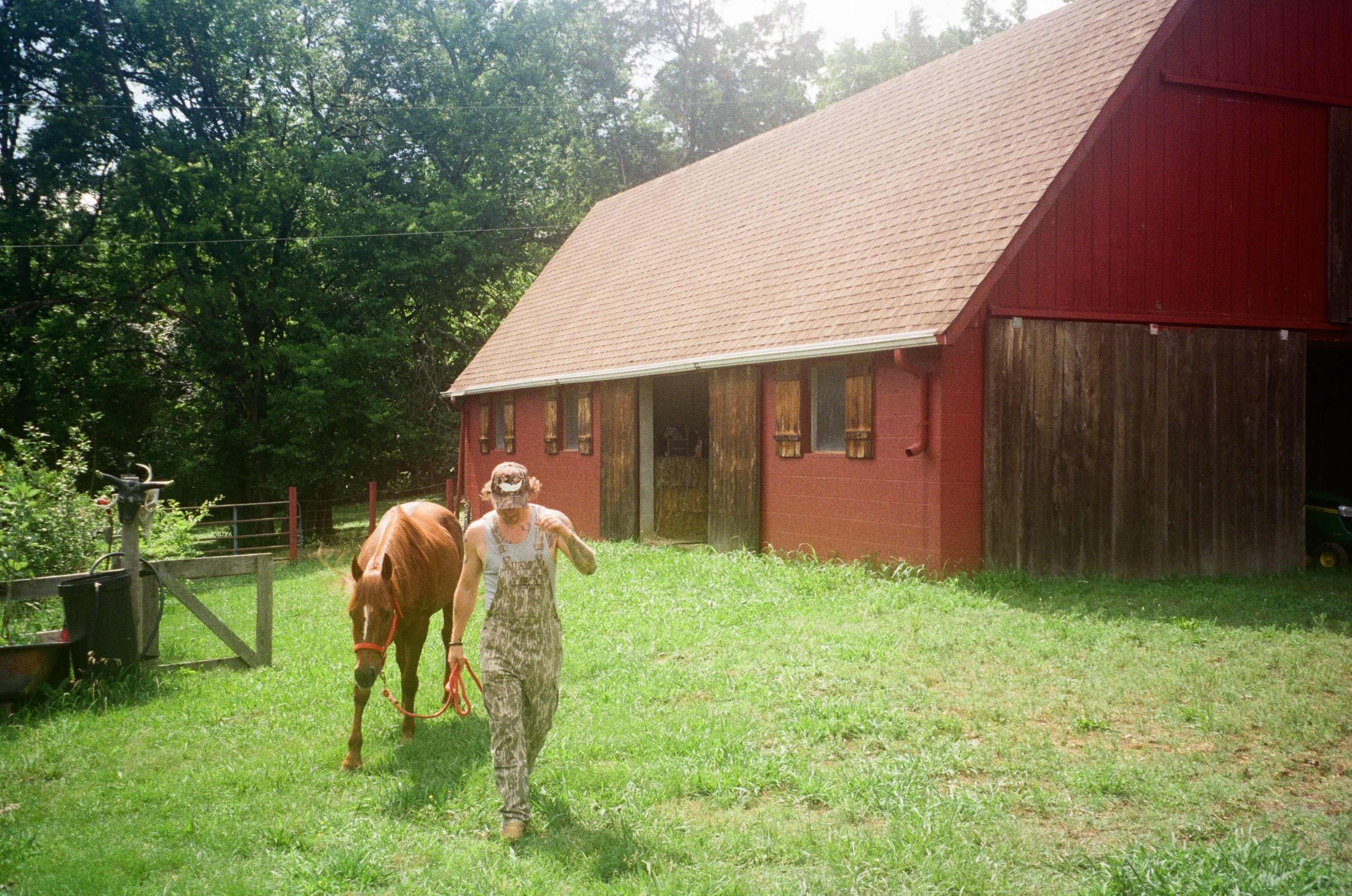 A person in camouflage overalls leads a chestnut horse towards a red barn surrounded by greenery.