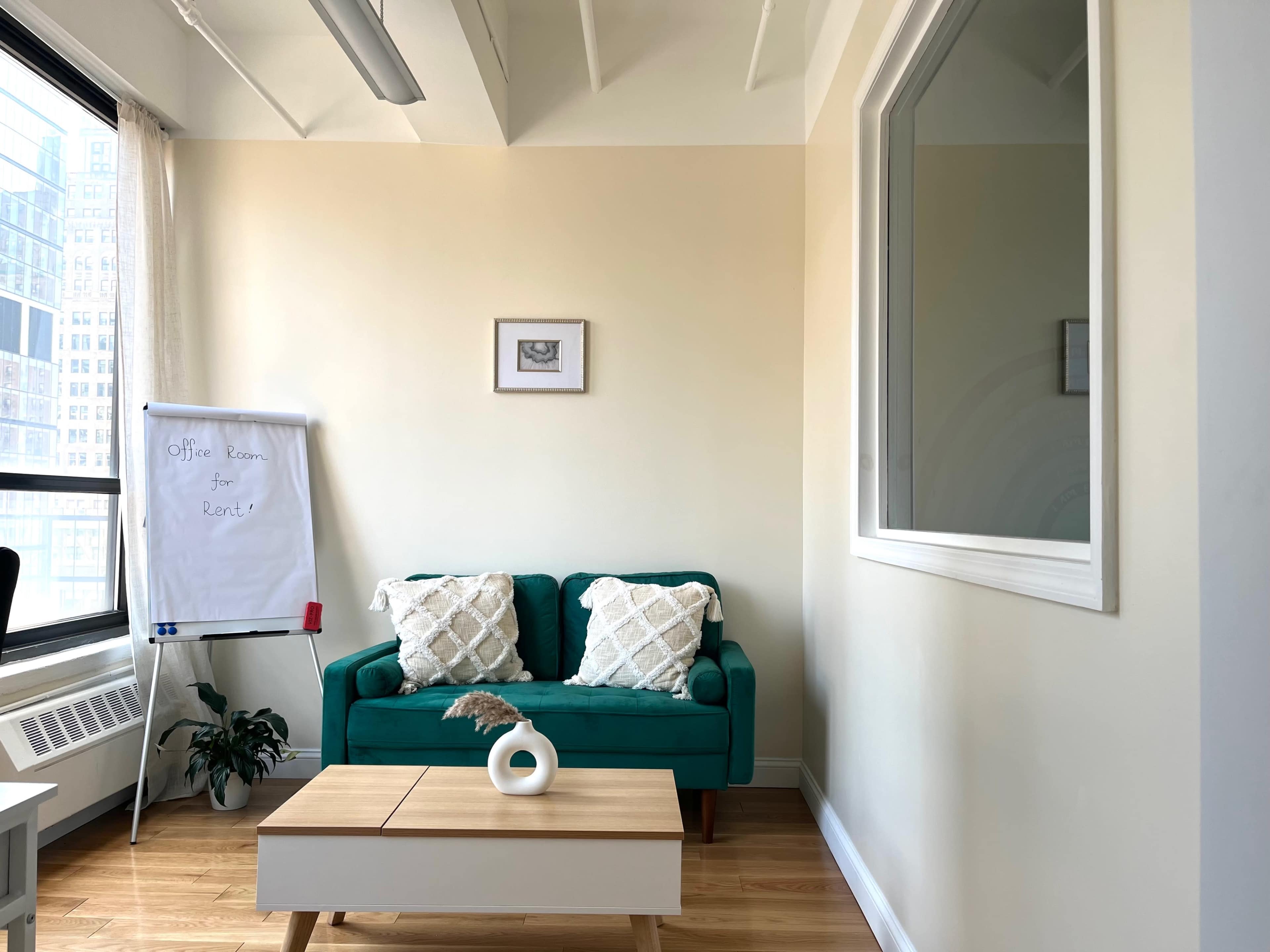 A bright office space featuring a teal sofa, a whiteboard with "Office Room for Rent" written on it, and a small coffee table with a vase, all positioned near a window.