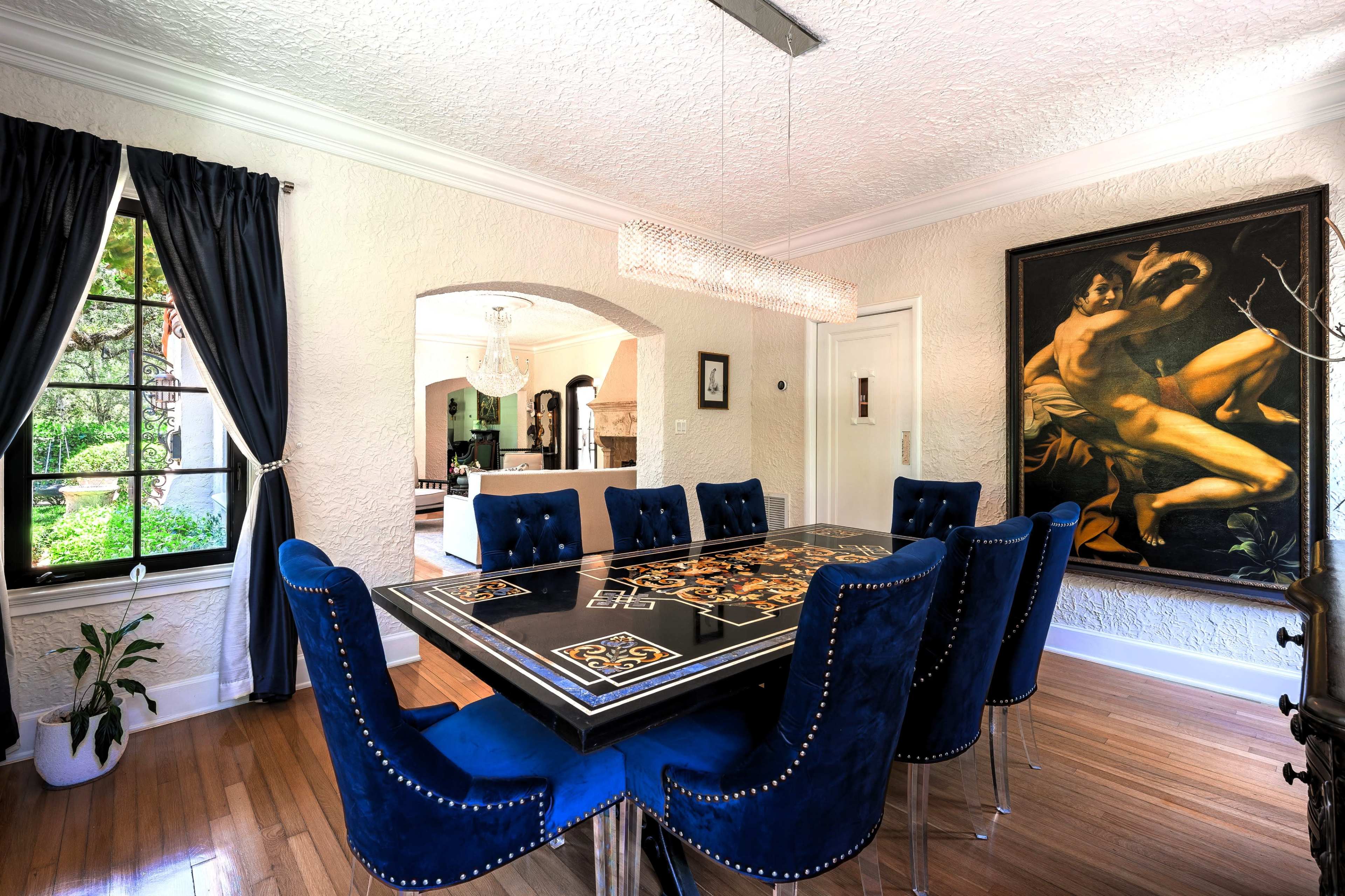 A dining room features a large, ornate black table surrounded by eight blue velvet chairs, with a chandelier overhead and a large painting on the wall.