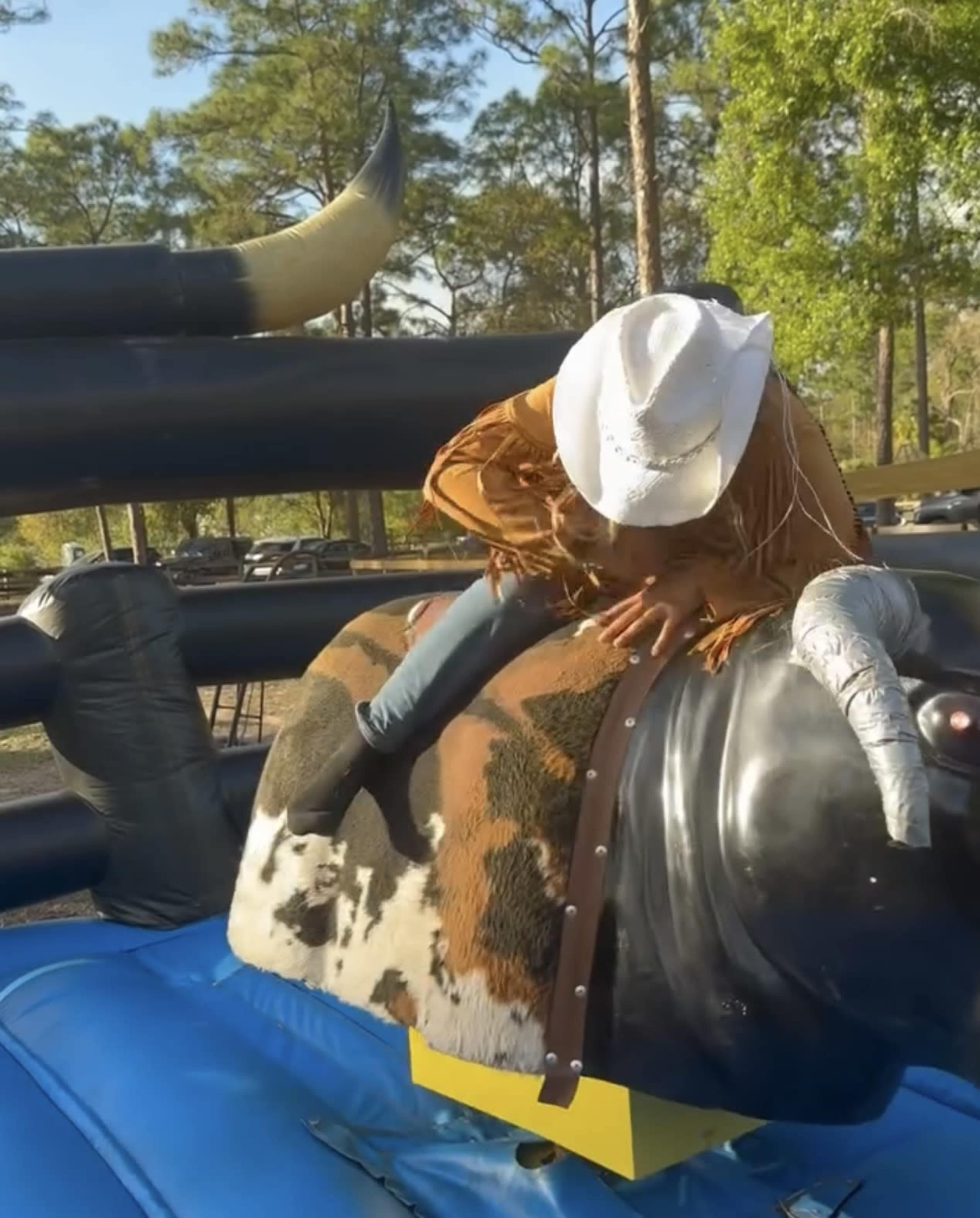 A person wearing a cowboy hat and fringed clothing is attempting to ride a mechanical bull at an outdoor event.