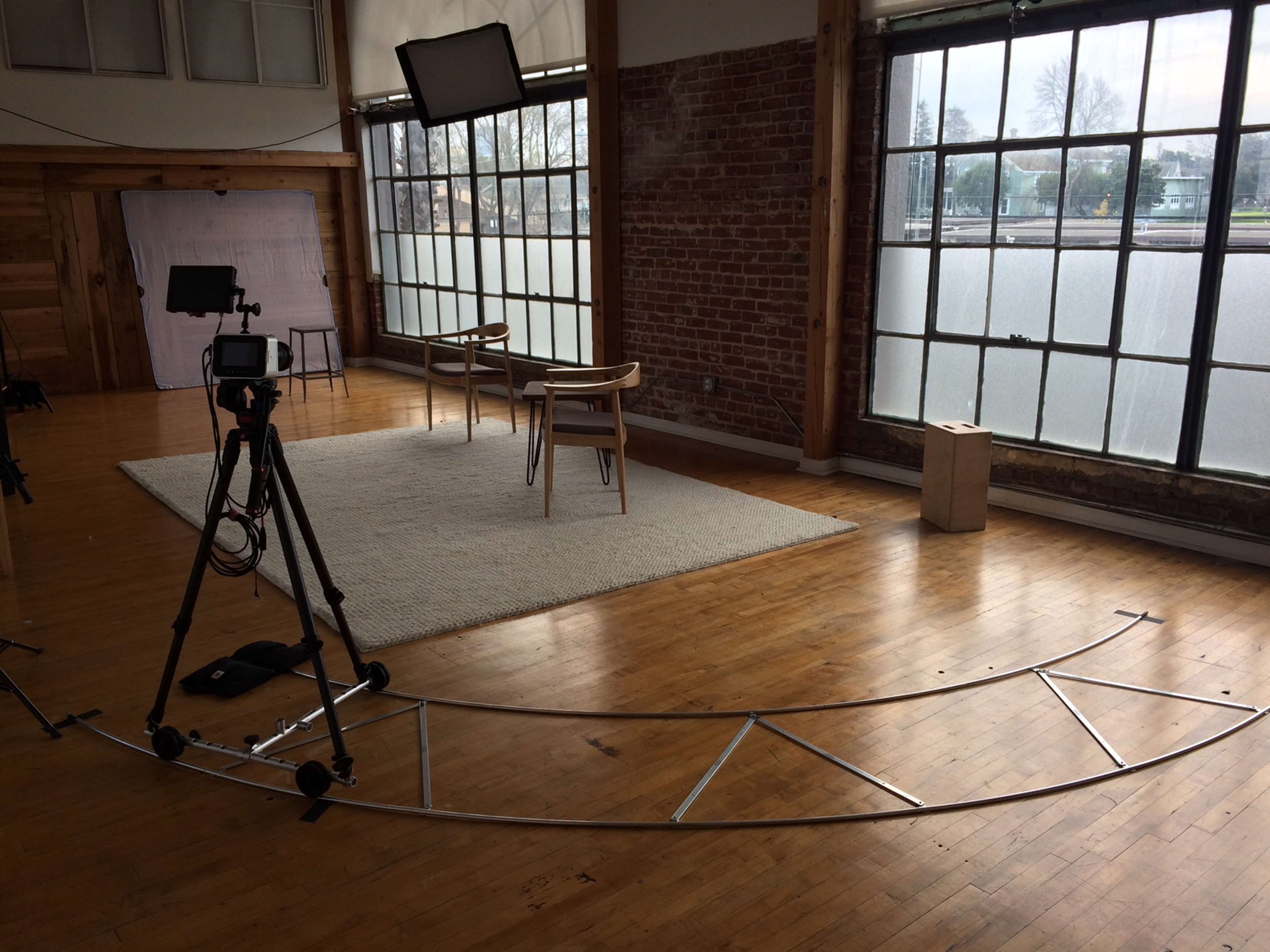 The image shows a film set featuring a circular camera track, a tripod with a camera, two chairs, and large windows letting in natural light.