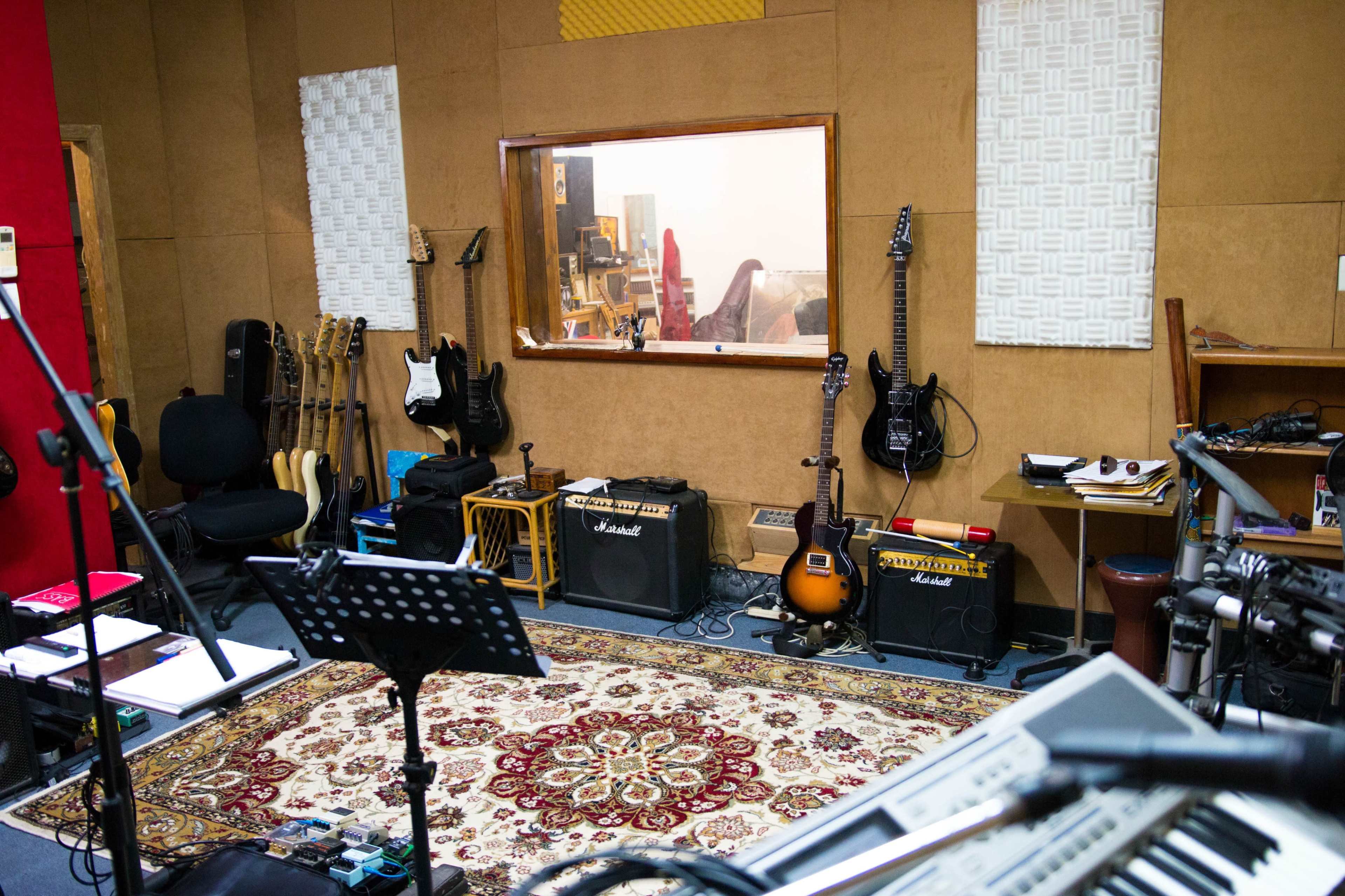 The image shows a music recording studio with various guitars, amplifiers, and a patterned rug, along with a control room visible through a window.