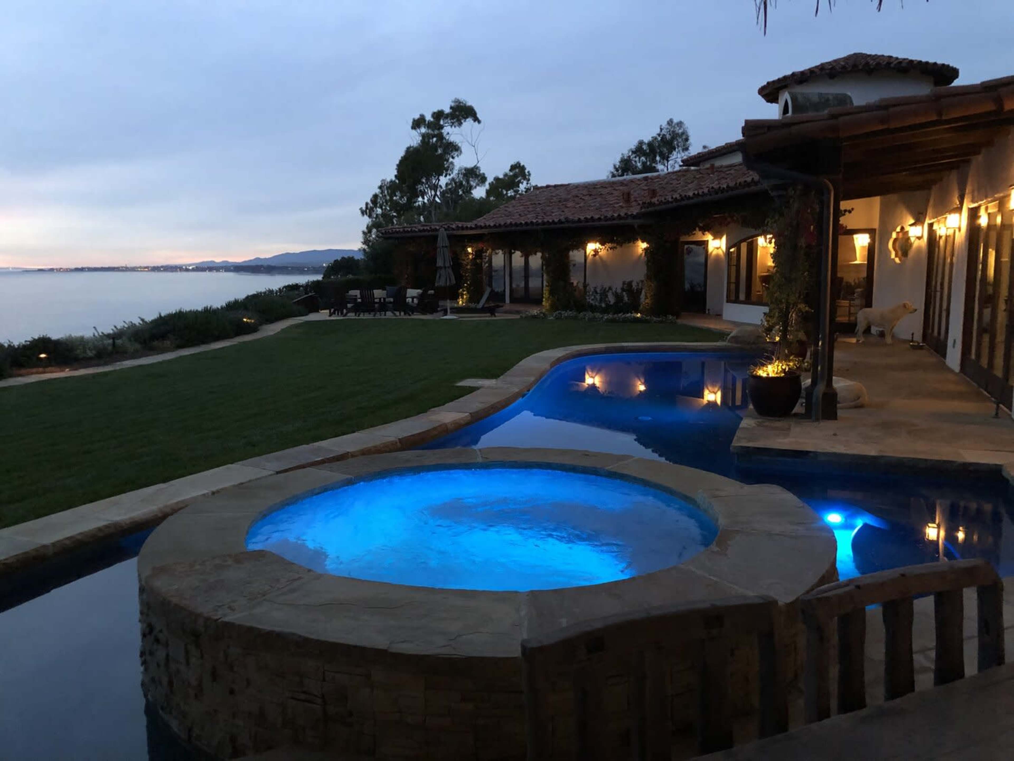 A backyard with a swimming pool and a spa, surrounded by grass and trees, facing a body of water under a cloudy sky.