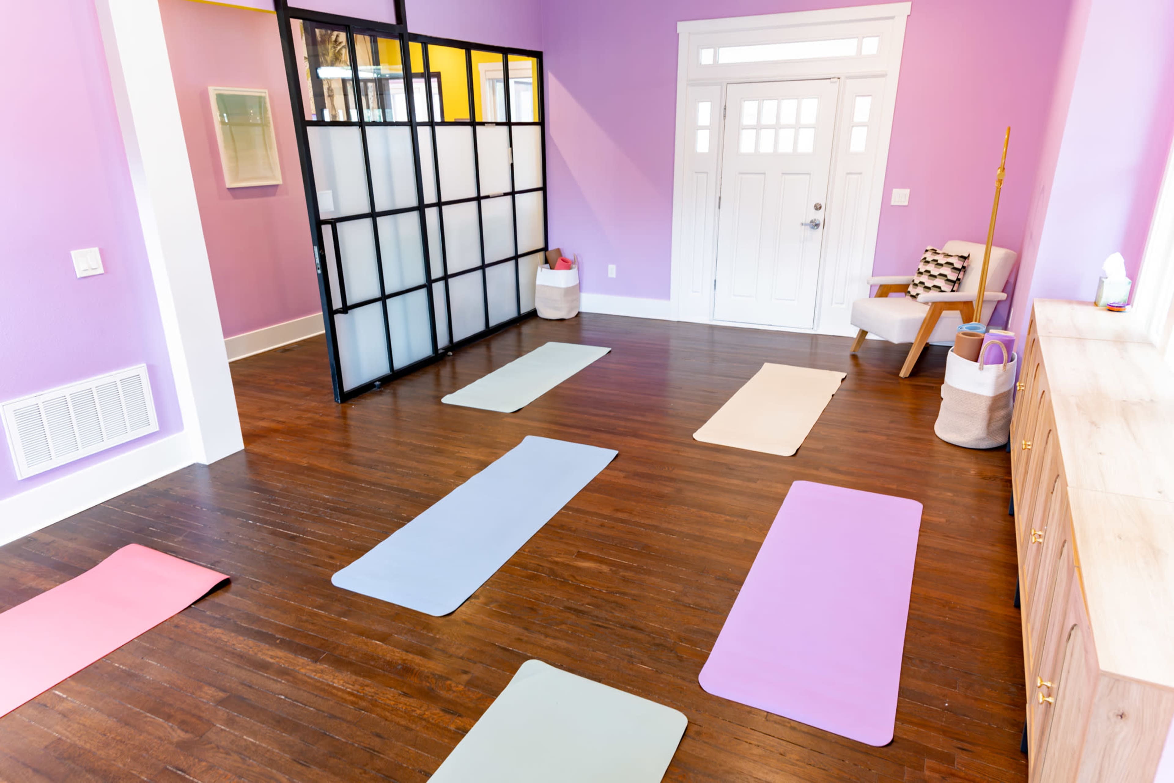 A spacious room features colorful yoga mats arranged on a wooden floor, with pastel-colored walls and natural light streaming through large windows.