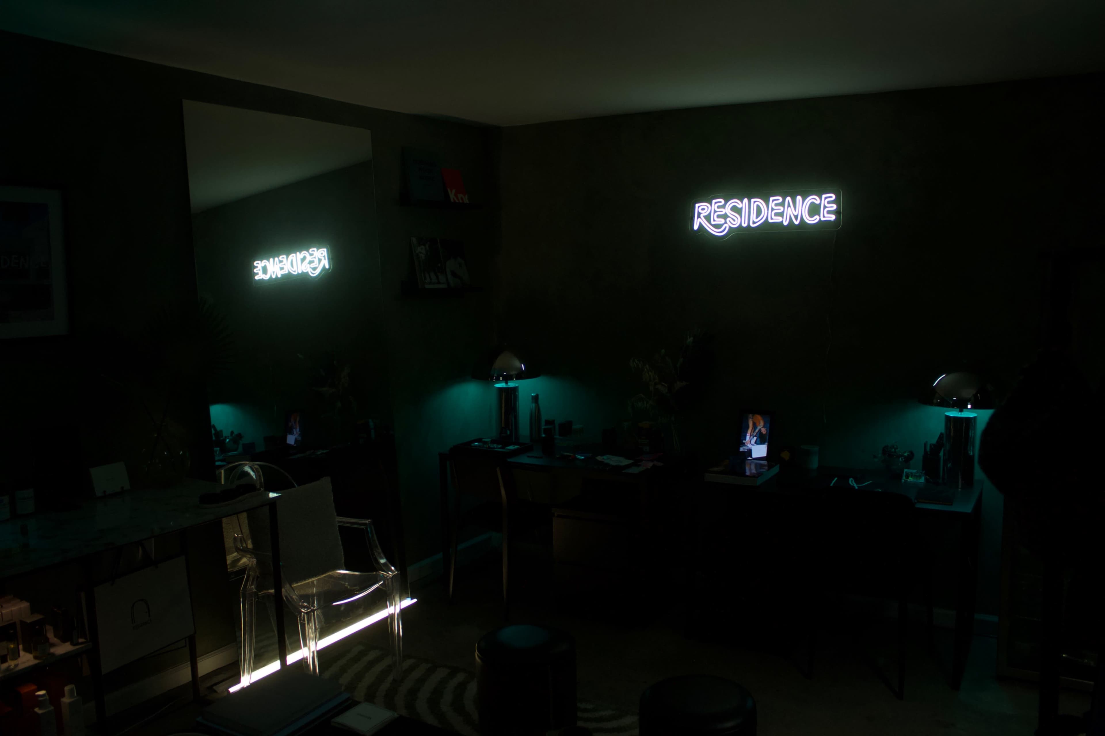 A dimly lit room features neon signs displaying the words "RESIDENCE" and "ENDEAVOR," with a reflection visible in a mirror and modern furniture arranged around the space.