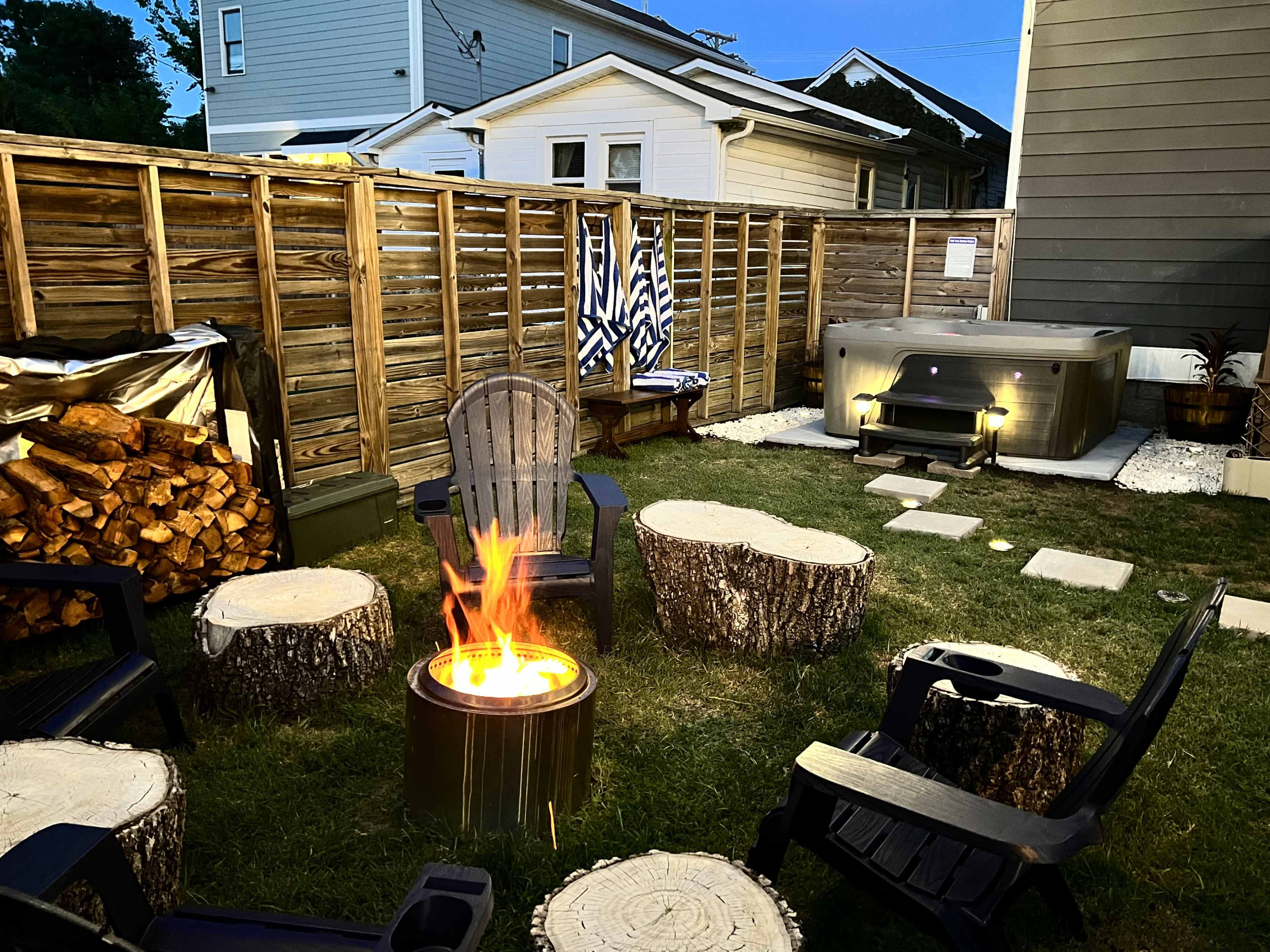 A backyard with a fire pit surrounded by wooden stump chairs, a hot tub in the corner, and a fenced area with stacked wood and a few decorative items.