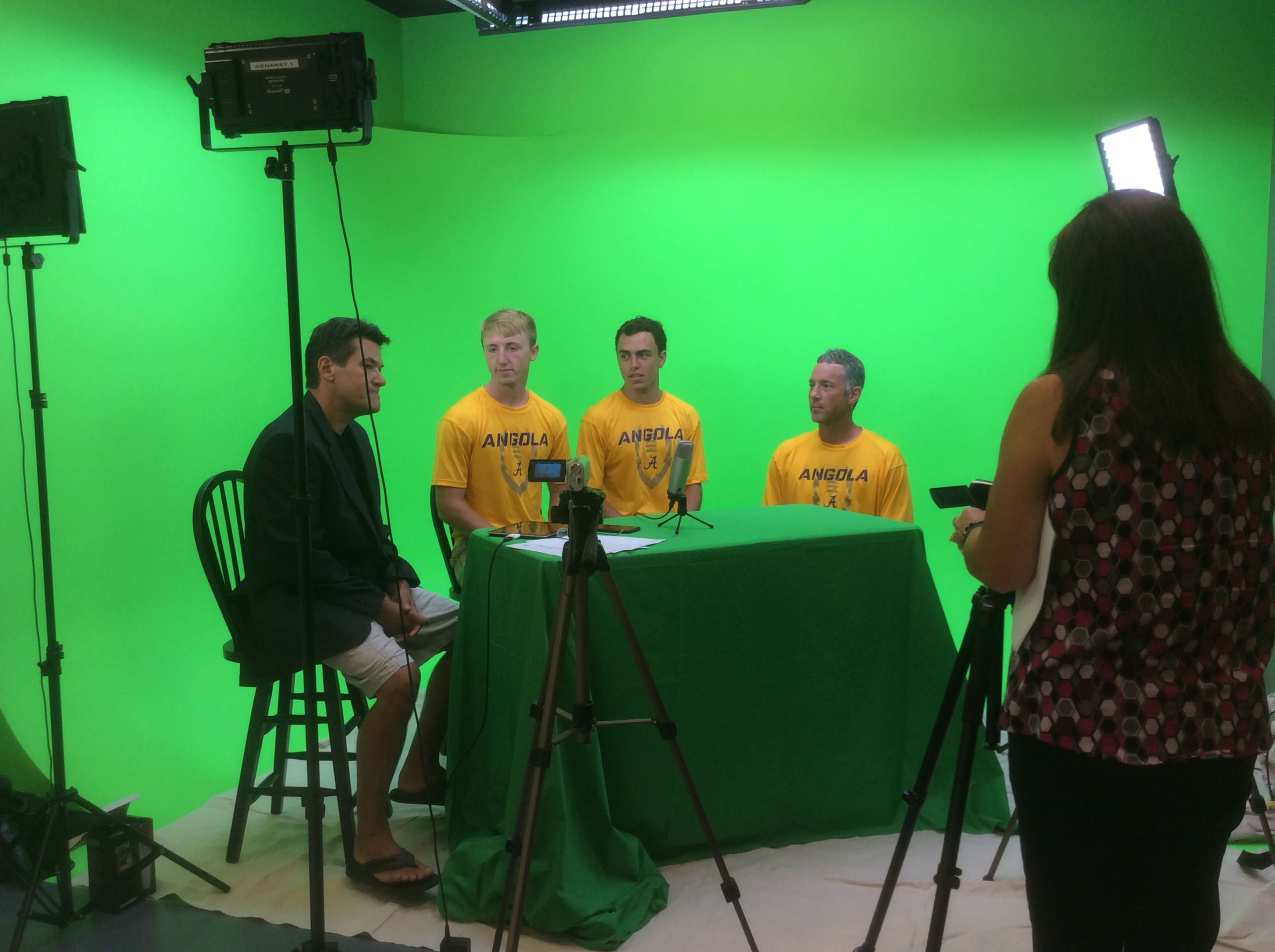 A group of four individuals, wearing yellow shirts marked "ANGOLA," is seated in front of a green screen, while a person on the right captures the scene with a camera.