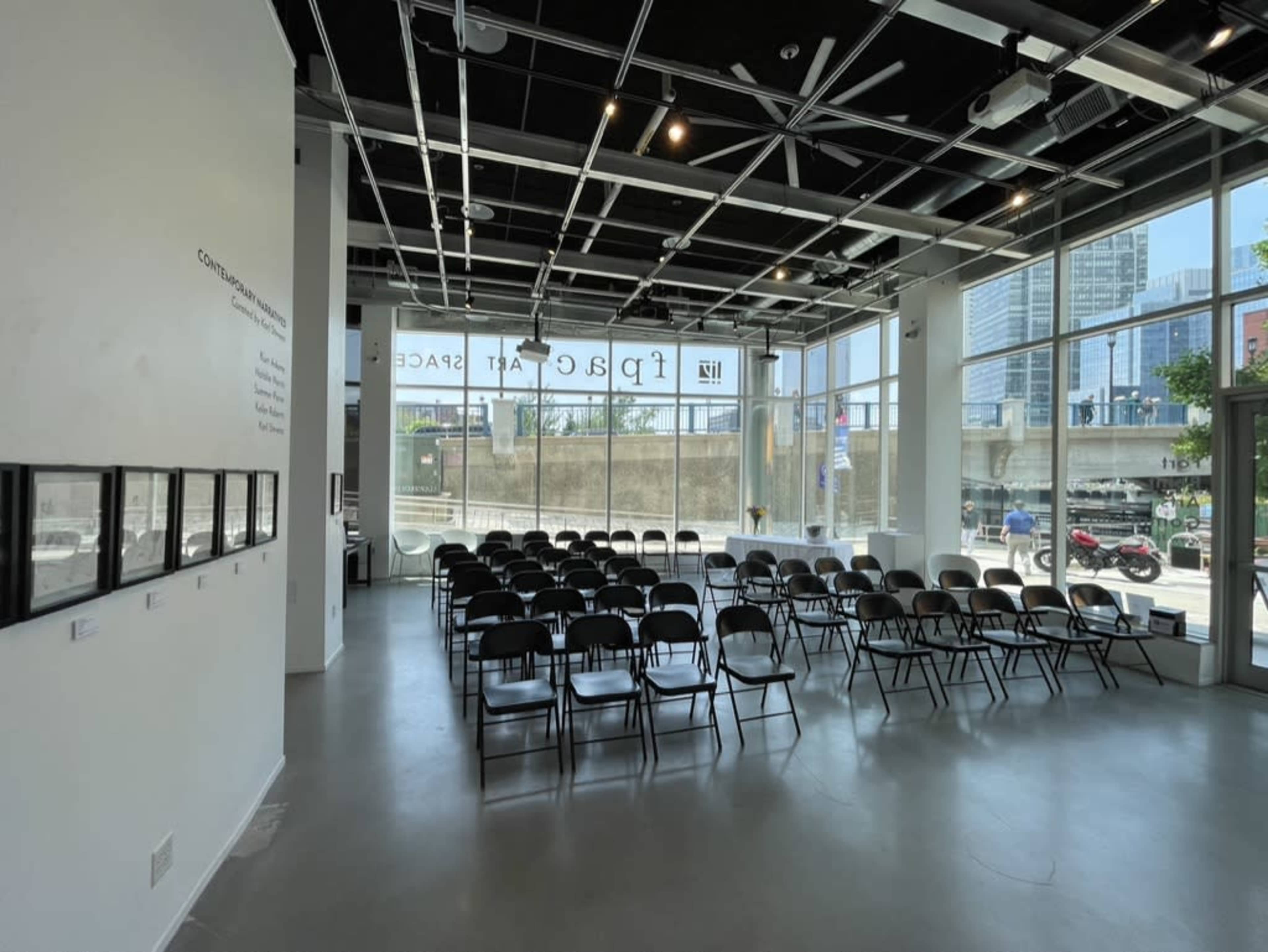 The image shows a modern, well-lit art space with rows of black chairs set up for an event, featuring large windows that overlook a cityscape.