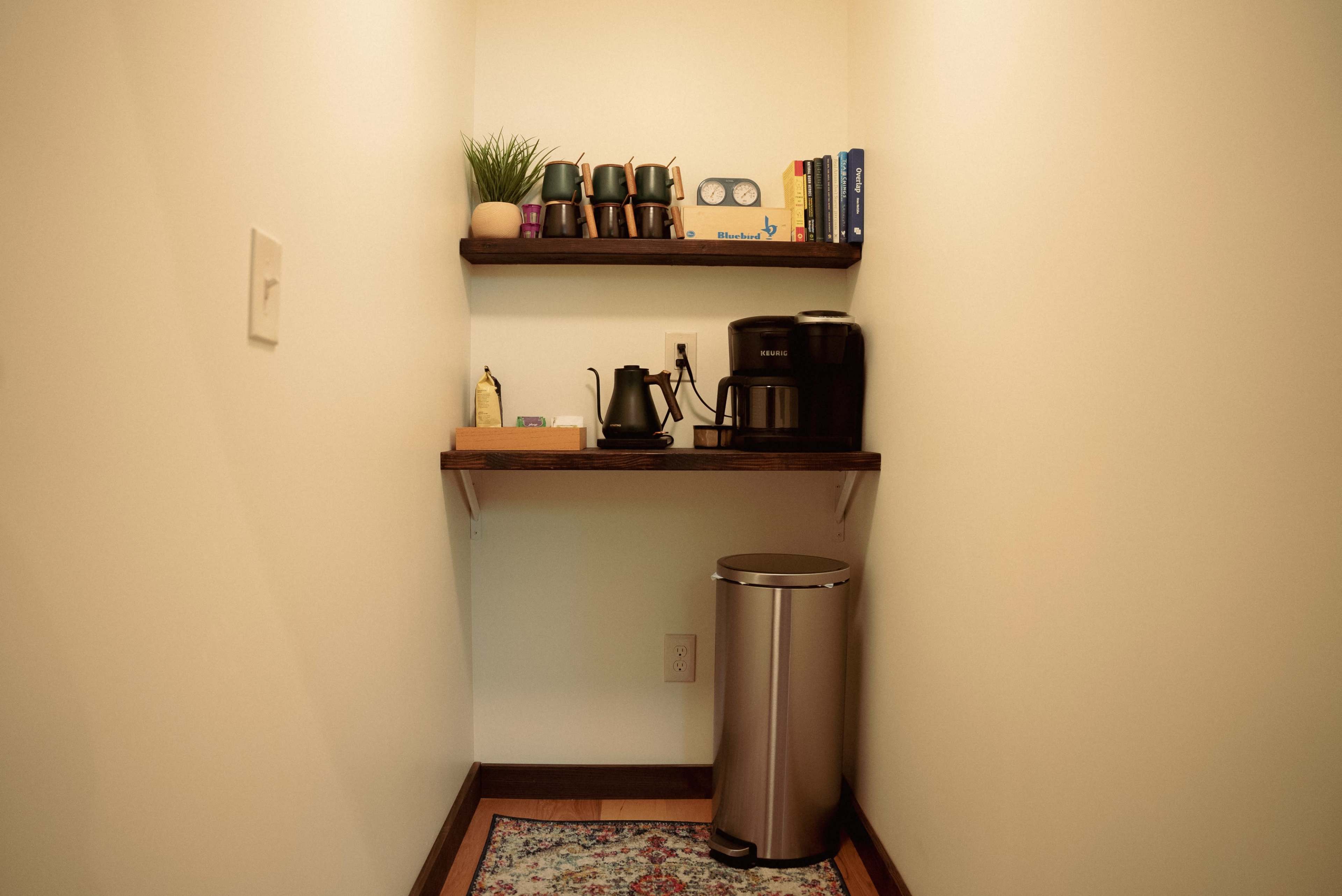 A small nook features a coffee station with shelves holding cups and books, alongside a coffee maker and trash can.