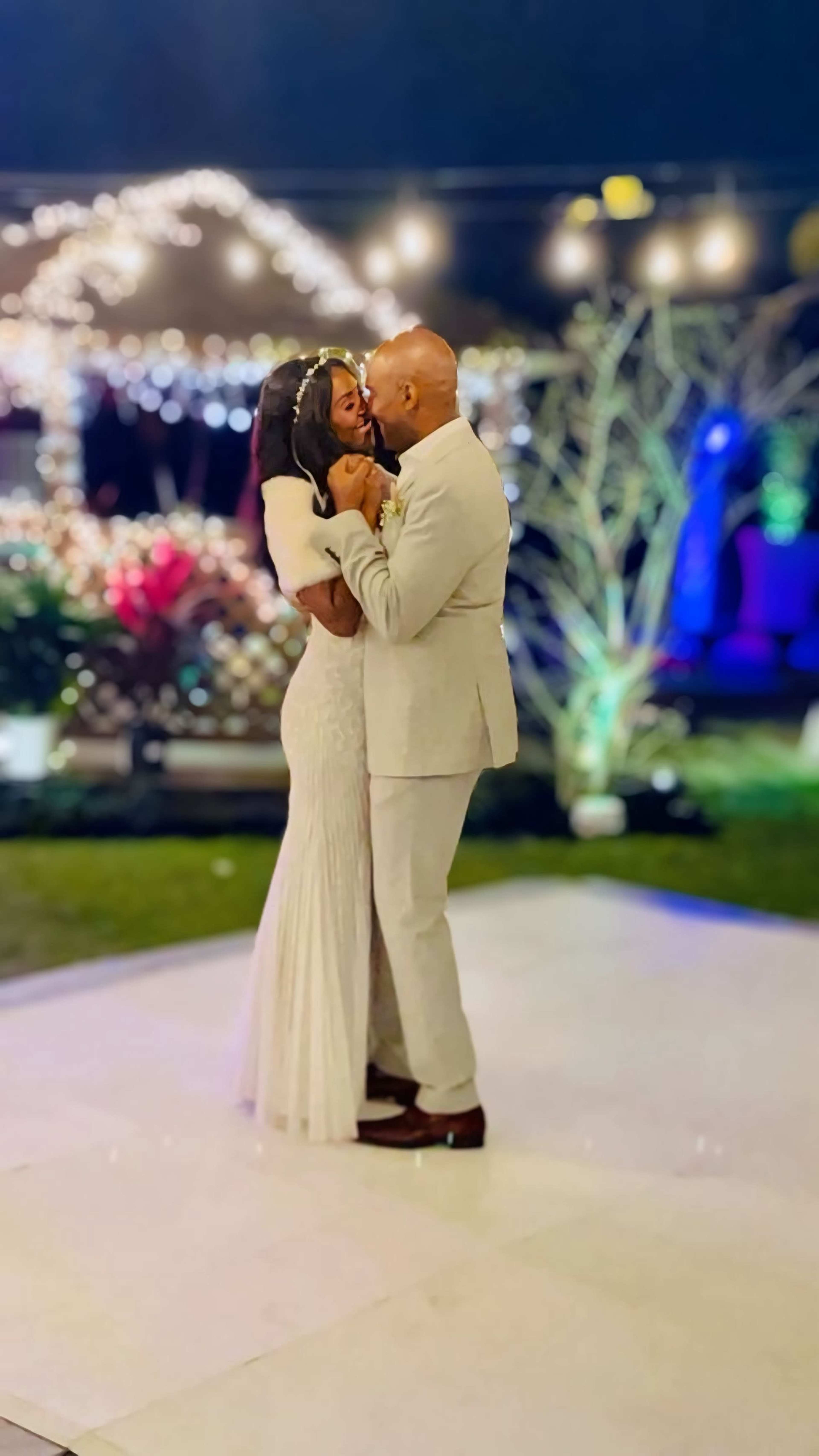 A bride and groom share a kiss while standing on a dance floor surrounded by festive lights and decorations.