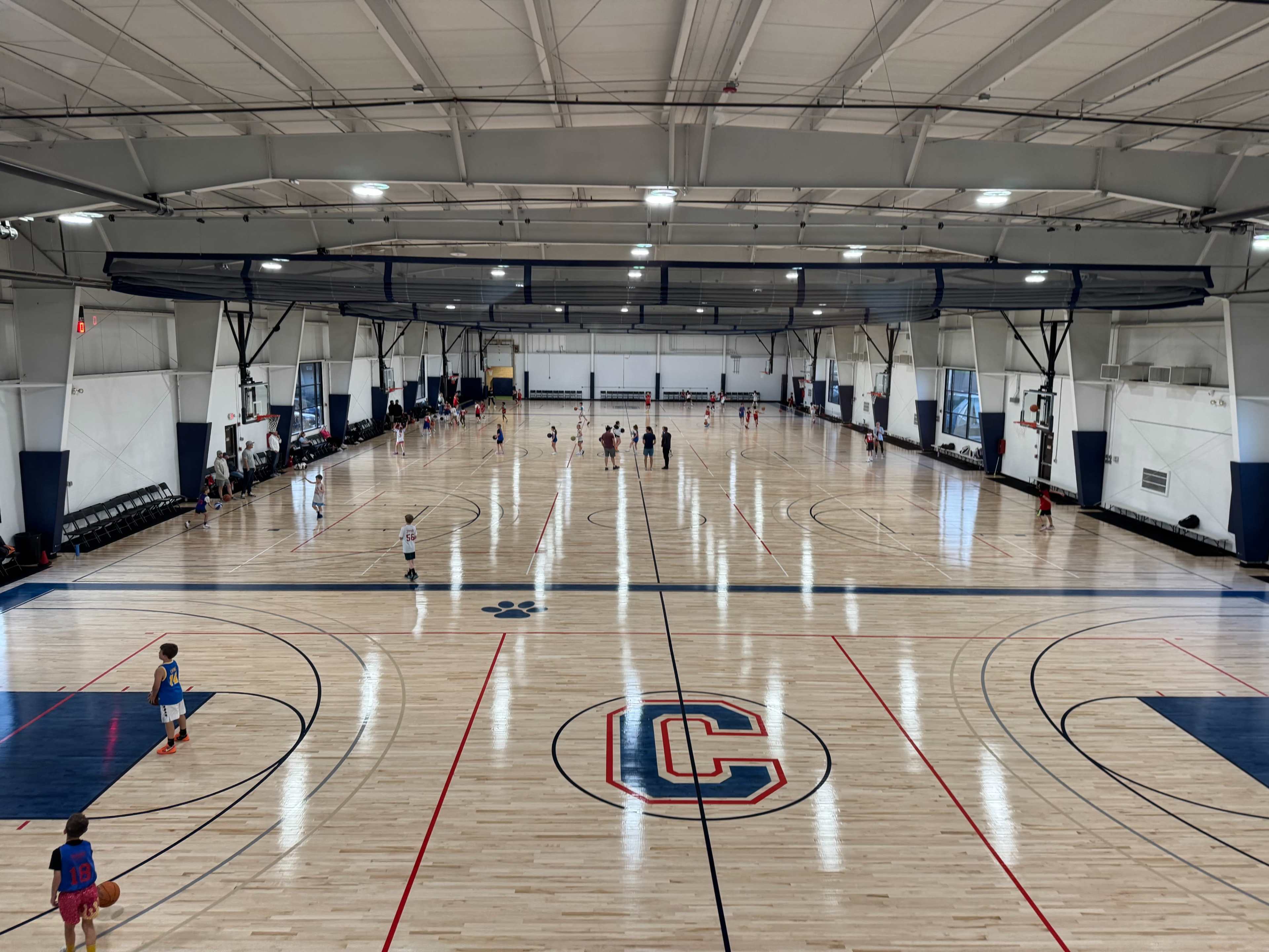 The image shows a large indoor basketball court with multiple playing areas, marked lines, and a central logo, while players are scattered throughout.