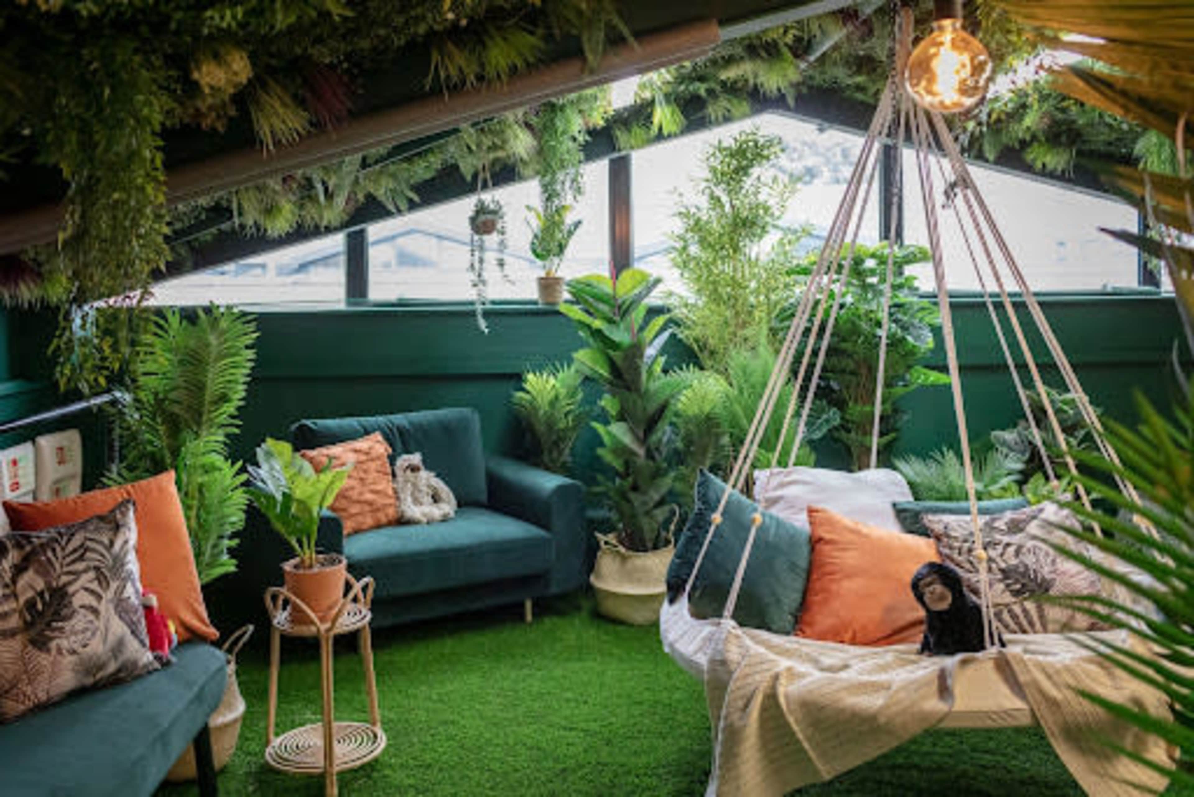 A cozy attic space filled with green plants, featuring a swing chair, a small side table, and two sofas with decorative cushions.