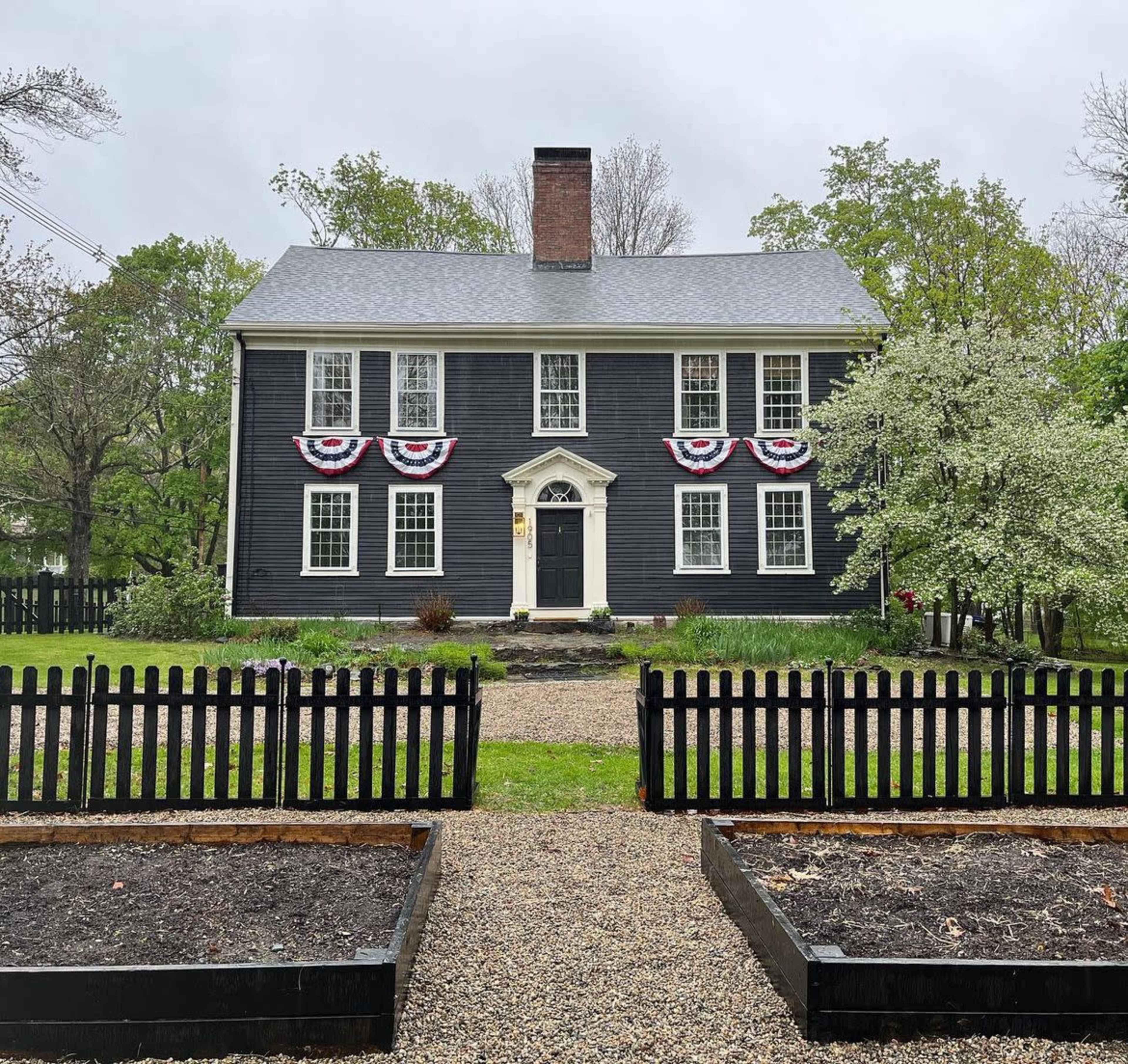 The image shows a two-story black colonial-style house with white trim, decorated with red, white, and blue bunting, surrounded by a picket fence and garden beds.