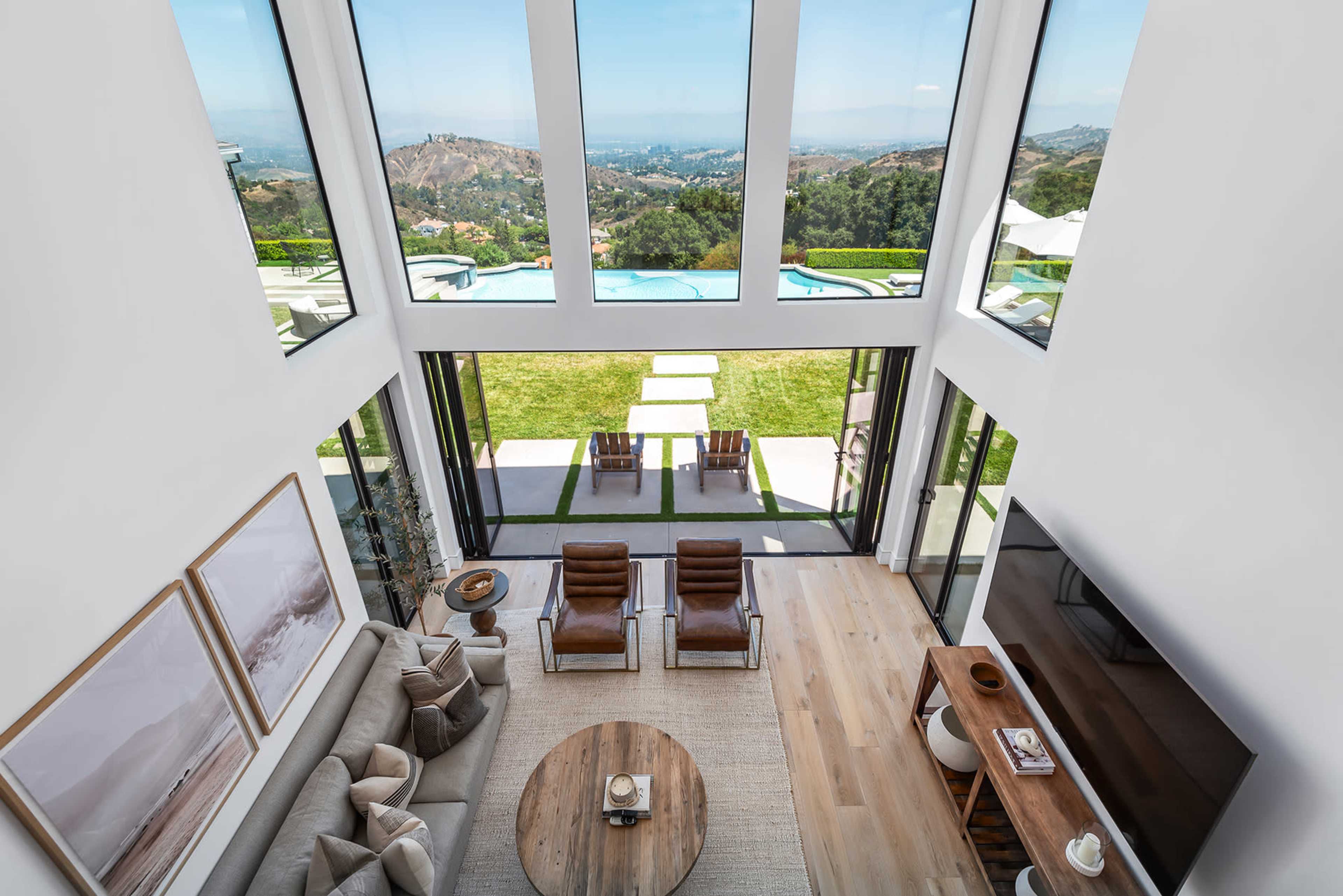 A modern living room with large windows offering a view of a landscaped yard and distant hills, furnished with a sofa, chairs, and a coffee table.