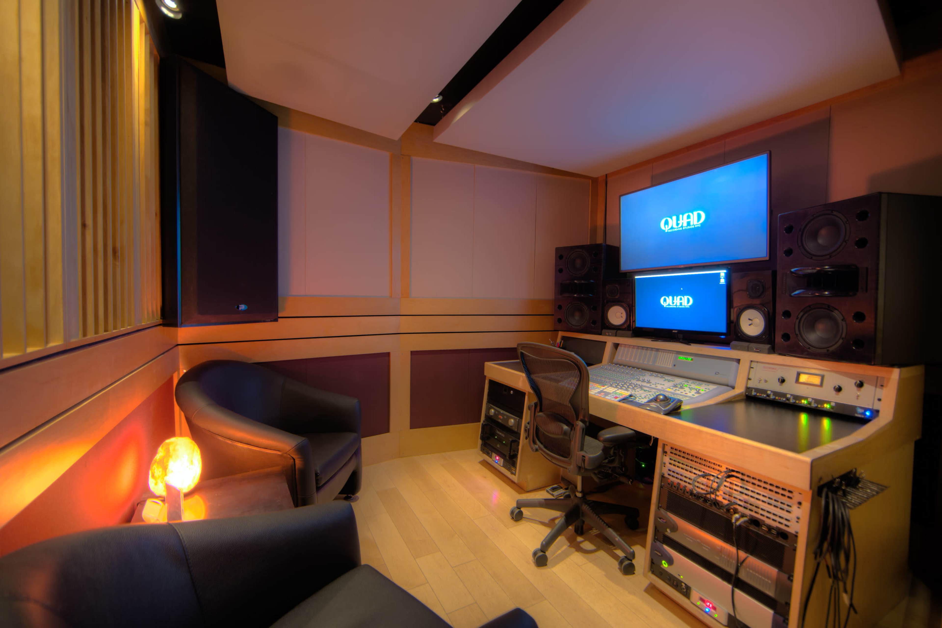 A recording studio featuring a control desk with audio equipment, large speakers, and a computer monitor, alongside two black chairs and a salt lamp in a cozy corner.
