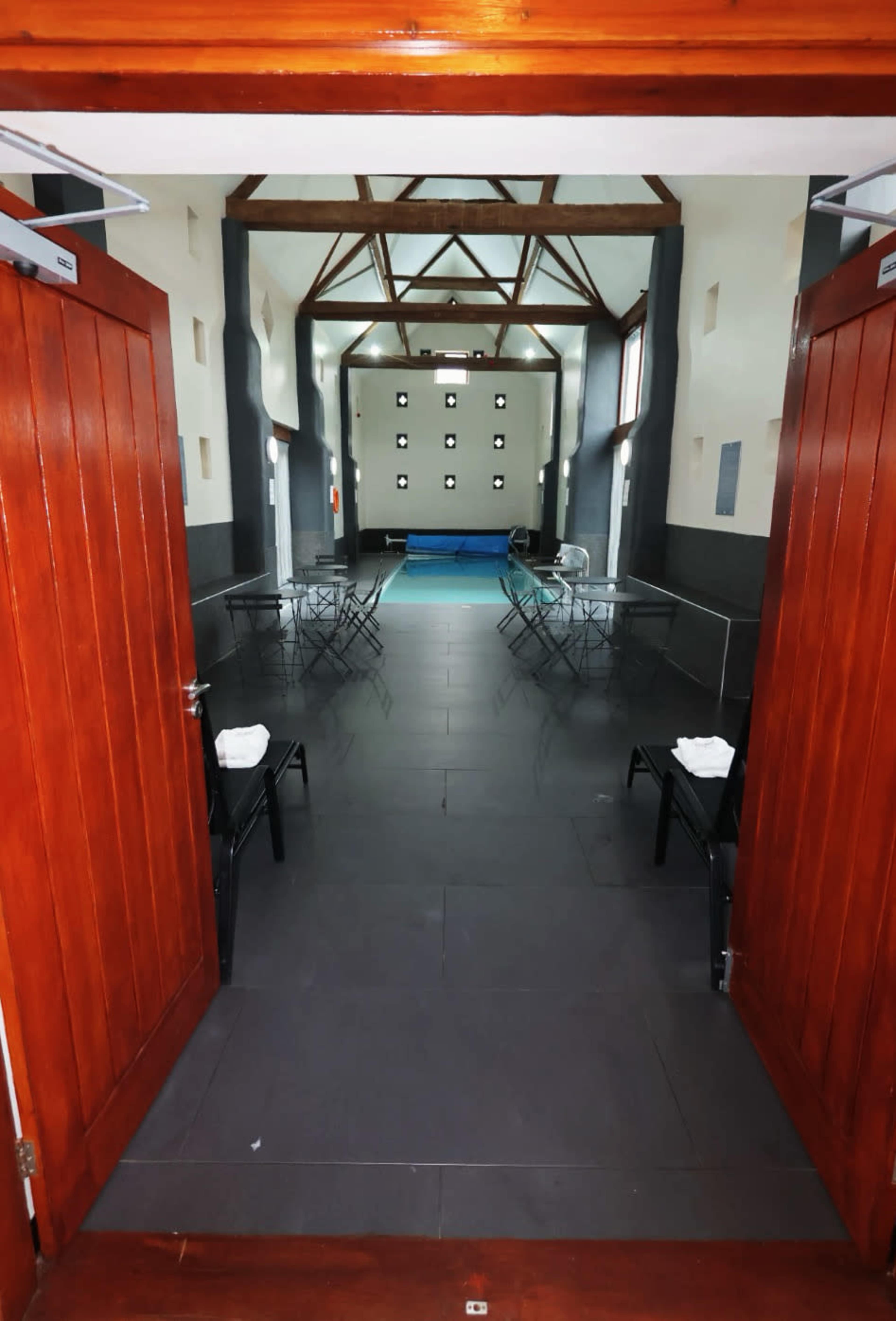 The image shows an interior space with two wooden doors open, revealing a long, spacious room with chairs arranged on either side and a pool at the far end.