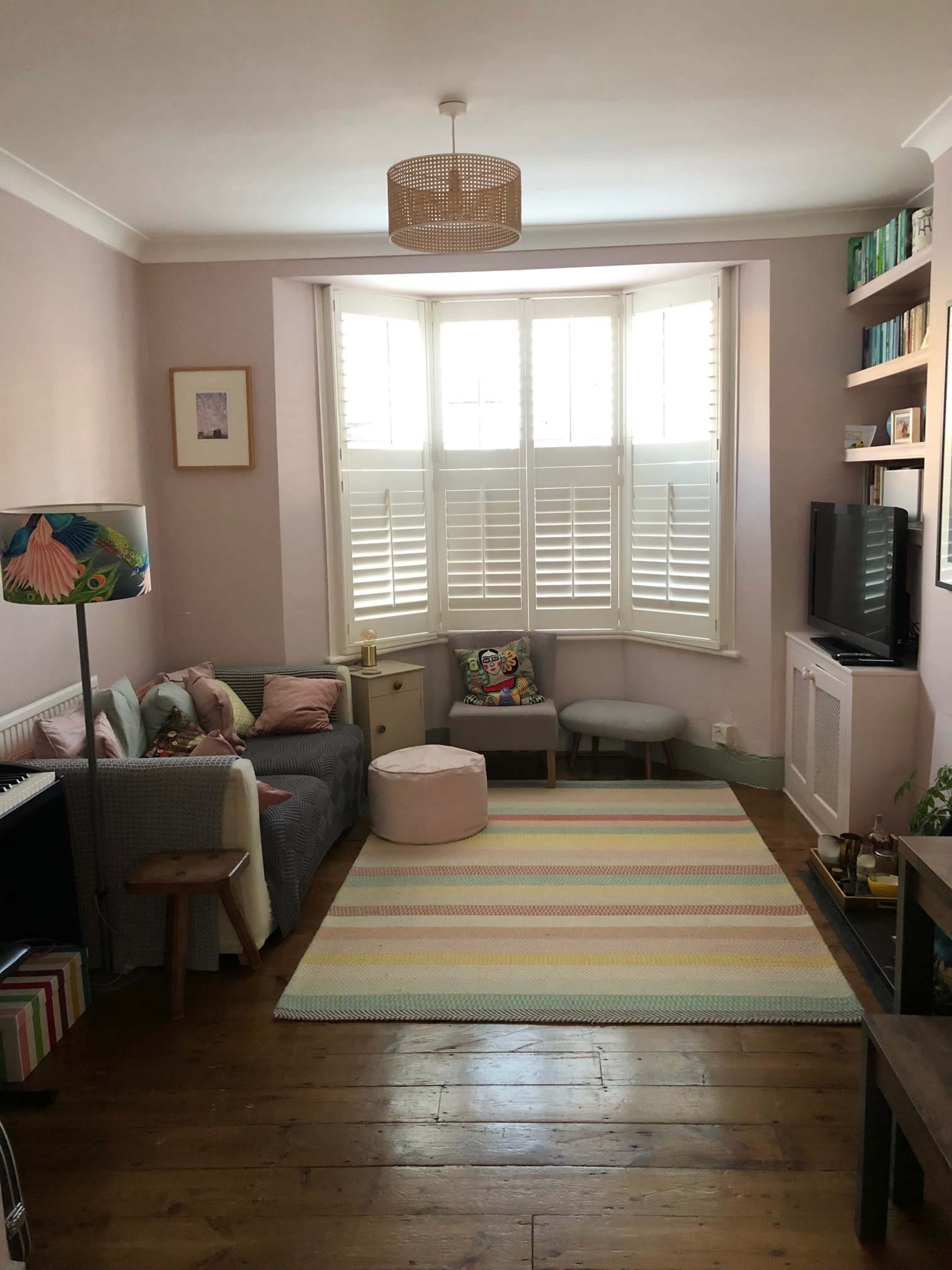 A living room with a light purple wall, a large bay window with shutters, a gray sofa, a round ottoman, and a striped rug on a wooden floor.