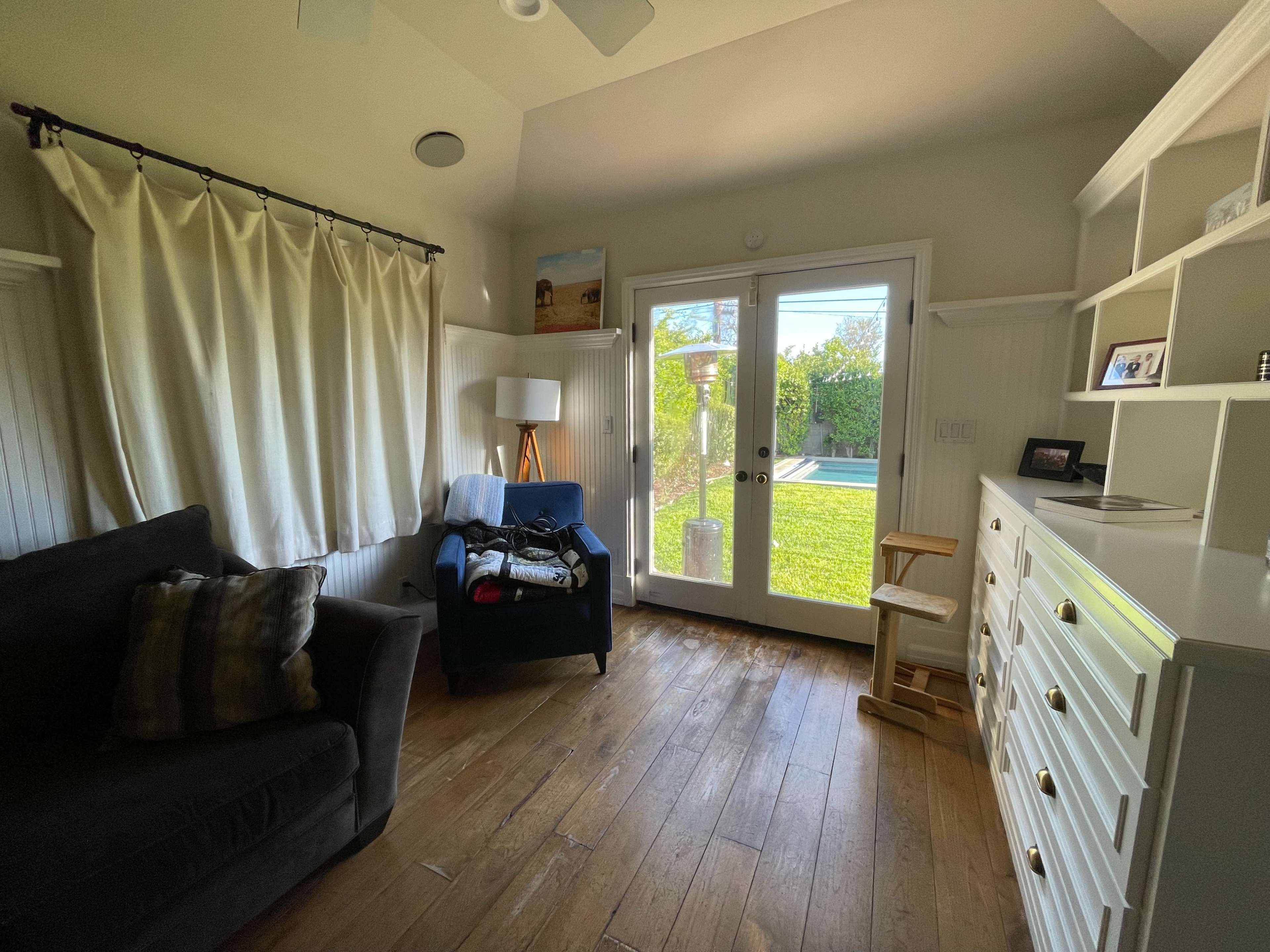 A cozy room with a dark sofa, a lamp, and white shelves, featuring a glass door that opens to a green yard.