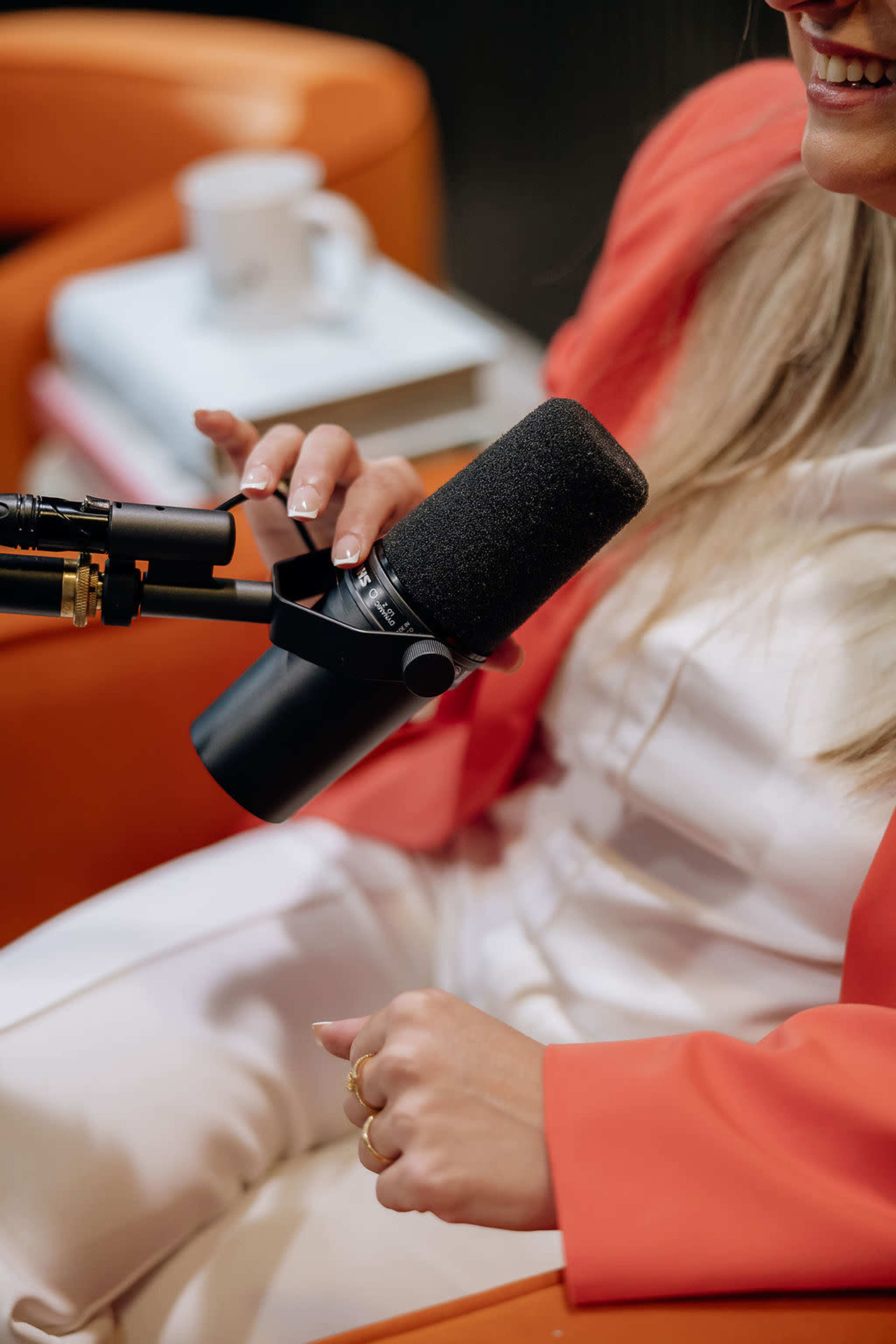 A woman in a coral blazer adjusts a microphone while sitting in a brightly colored armchair.