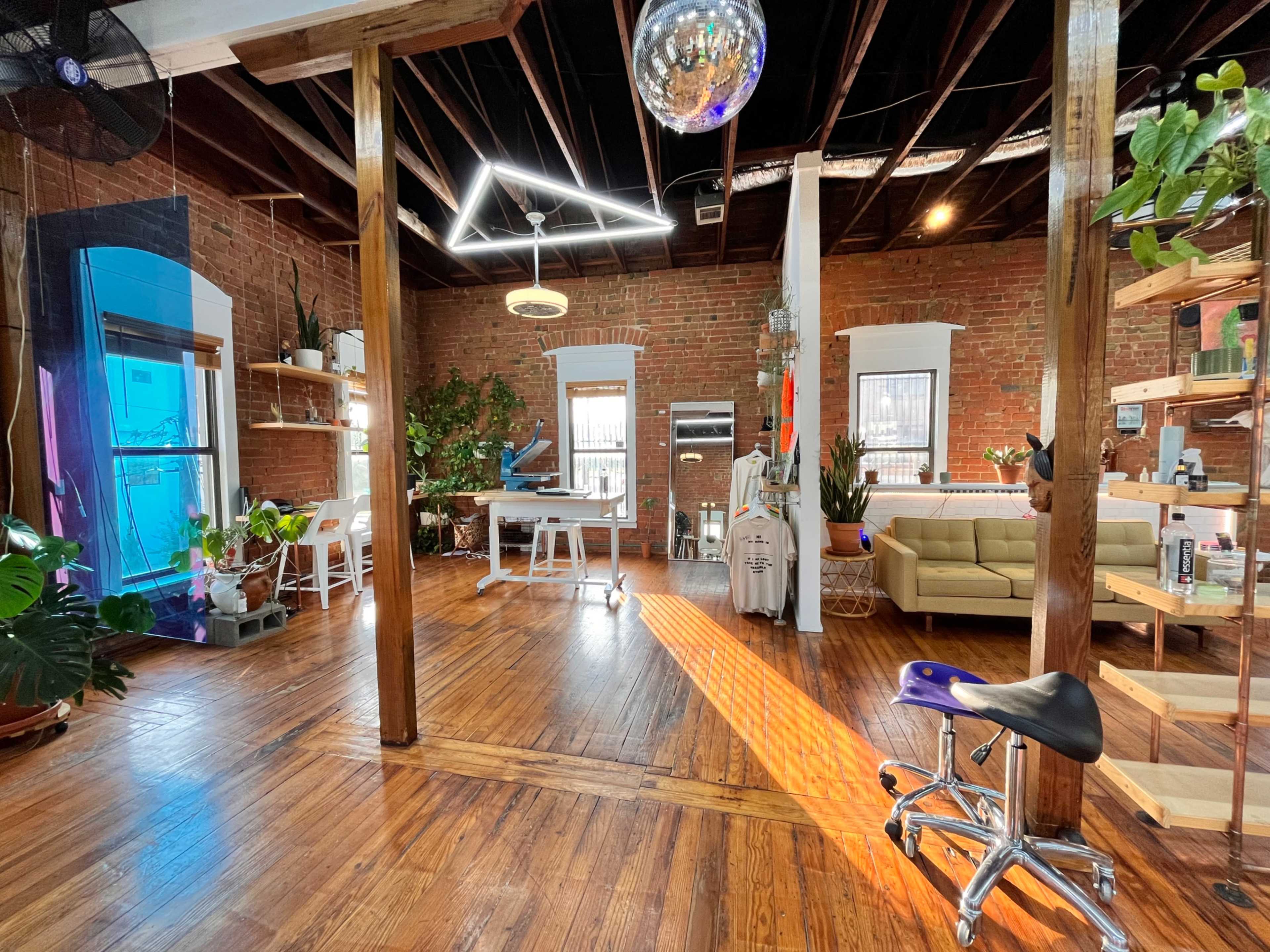 A spacious, well-lit studio with exposed brick walls, wooden flooring, and various plants, featuring a work desk, seating area, and a large mirror ball hanging from the ceiling.
