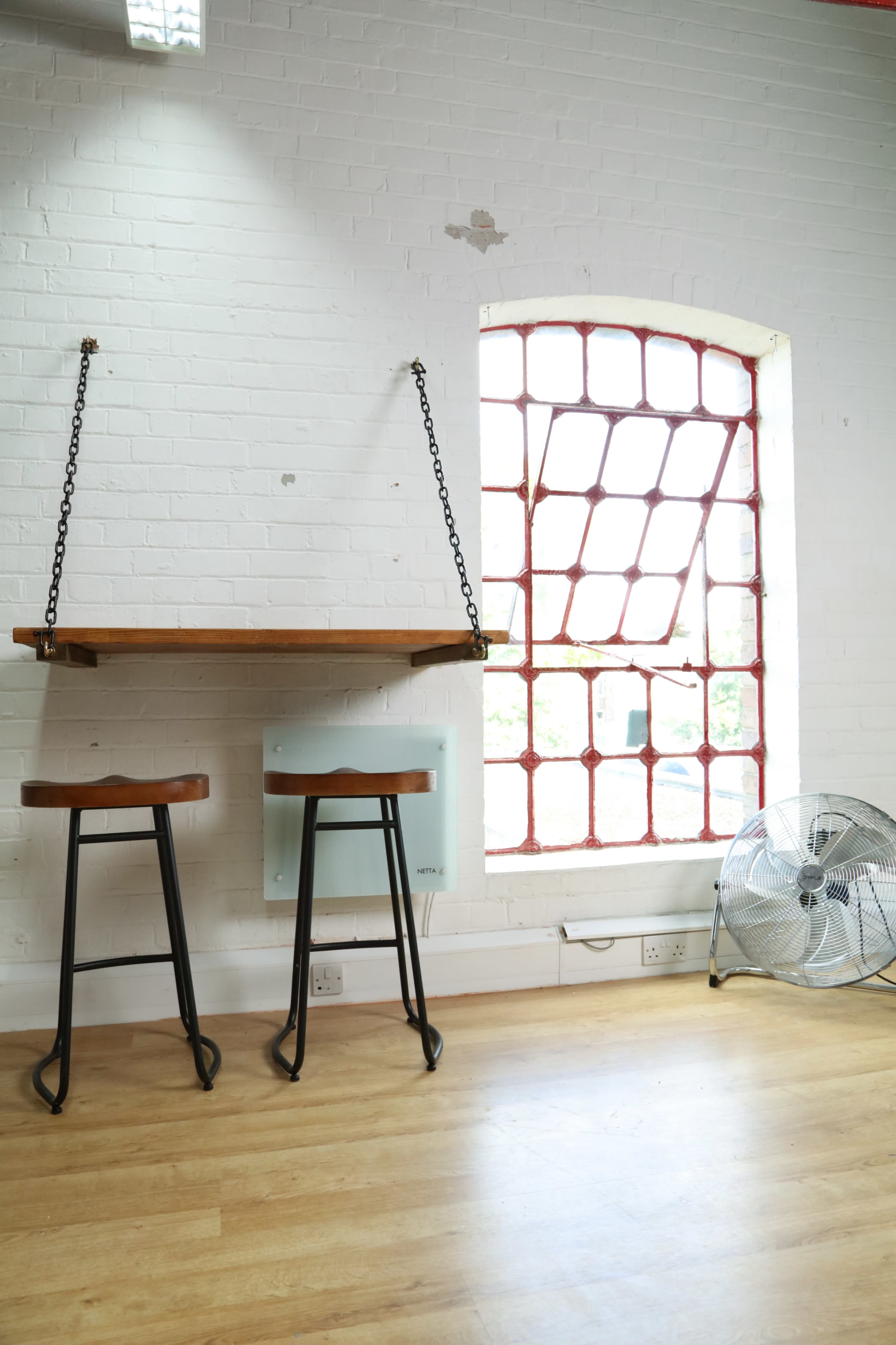 A minimalist space with a hanging wooden table, two metal bar stools, and a large red-framed window beside a floor fan.