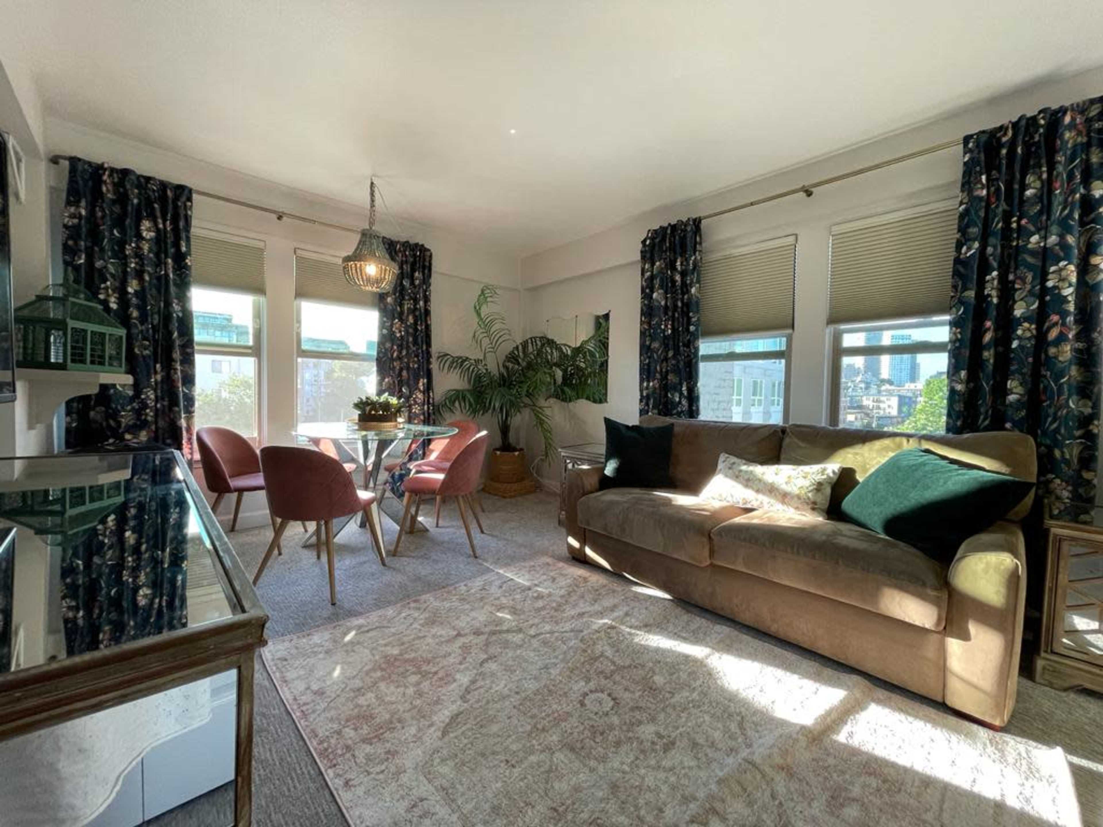 The image shows a bright living room with a sofa, a glass dining table surrounded by chairs, large windows, and plants in the corners.