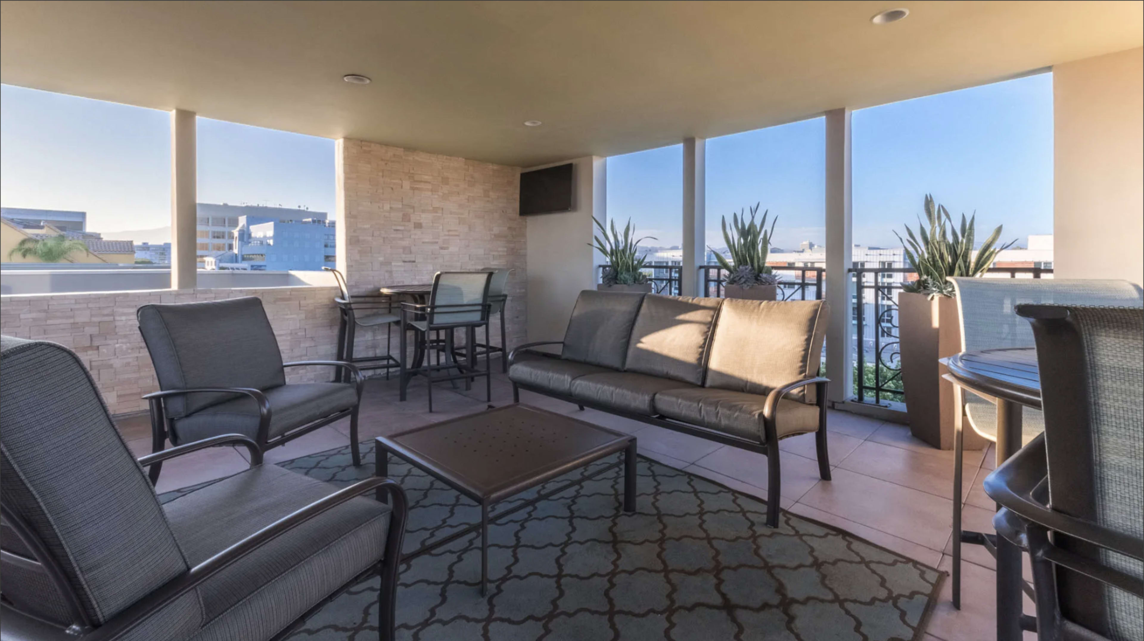 The image shows a modern balcony area featuring a seating arrangement with metal-framed chairs and a coffee table, along with a small dining table in the background, against a backdrop of clear skies.