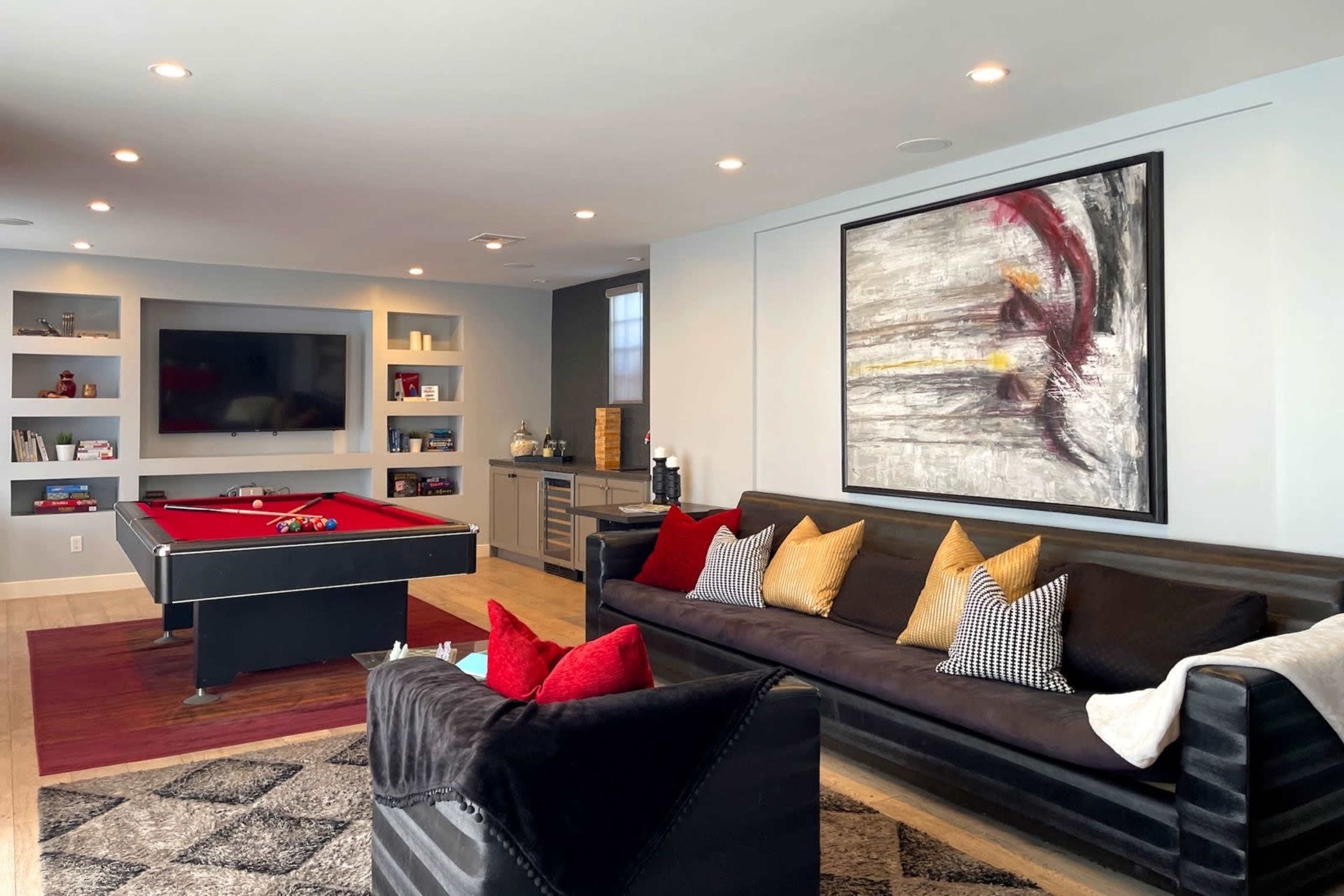 A modern game room features a red pool table, a black sectional sofa with decorative pillows, and a mounted television on the wall.