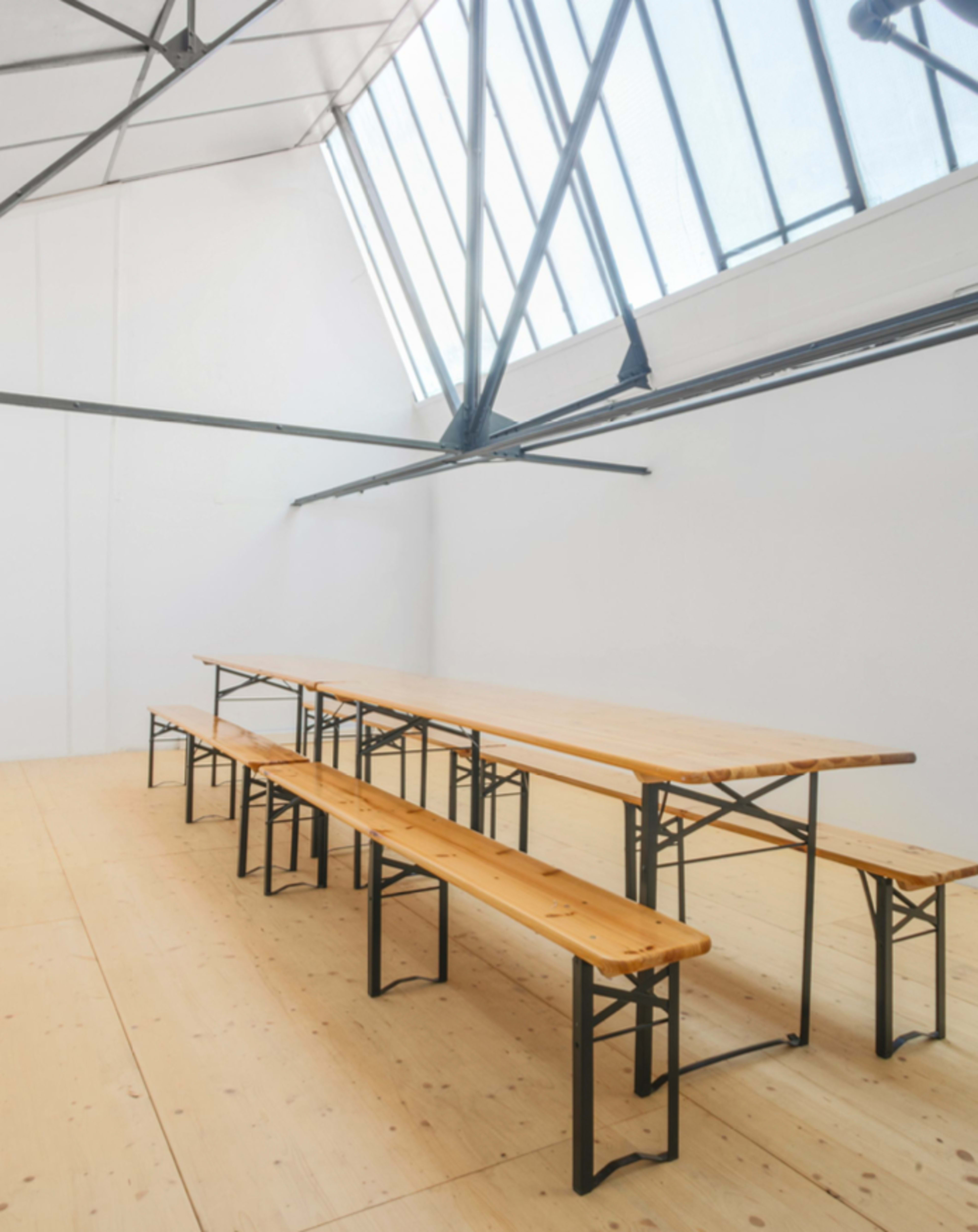 A spacious room features a long wooden table with matching benches and large windows allowing natural light to enter.