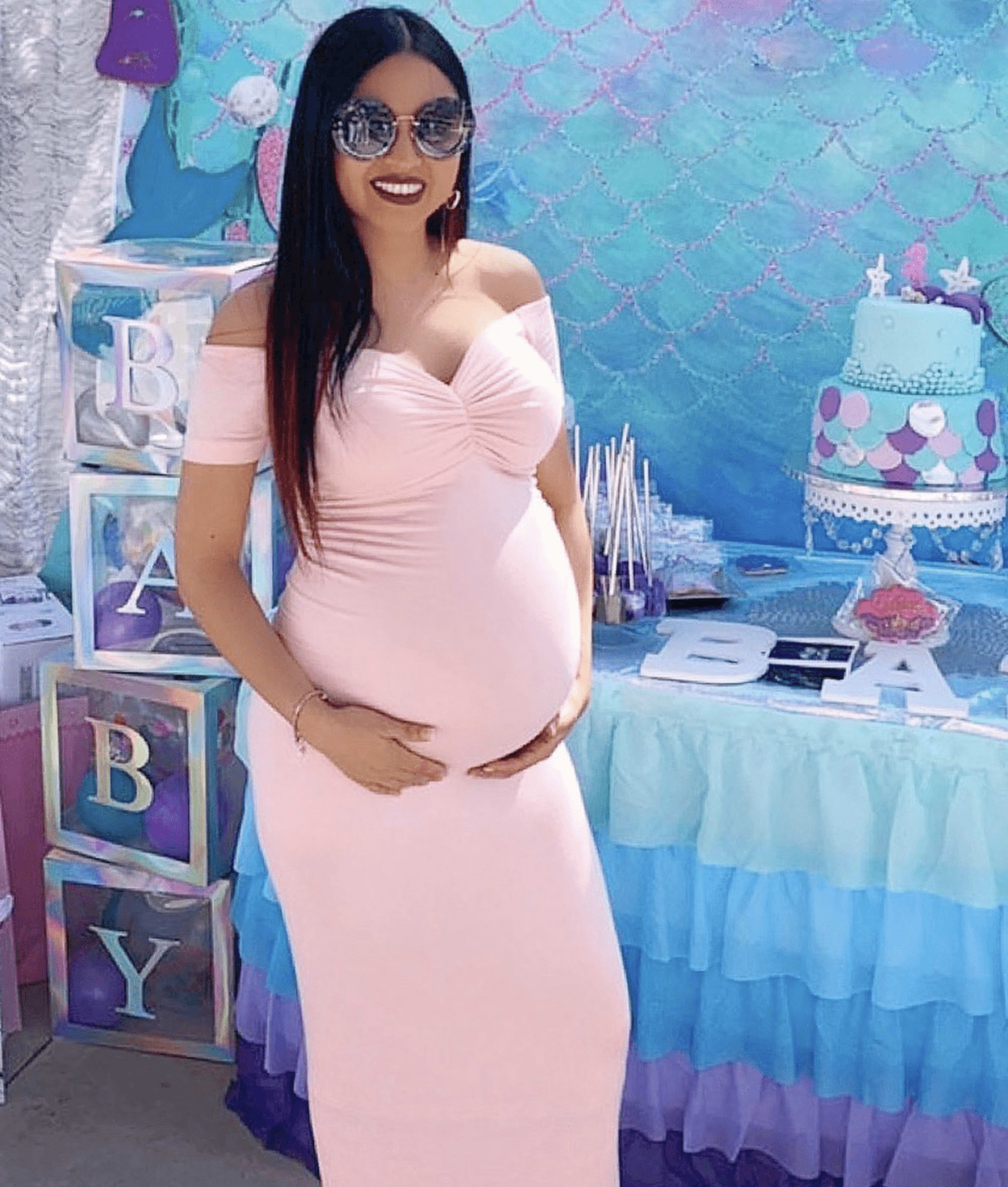 A pregnant woman in a pink dress poses in front of a decorated table featuring a cake and various baby-themed items.