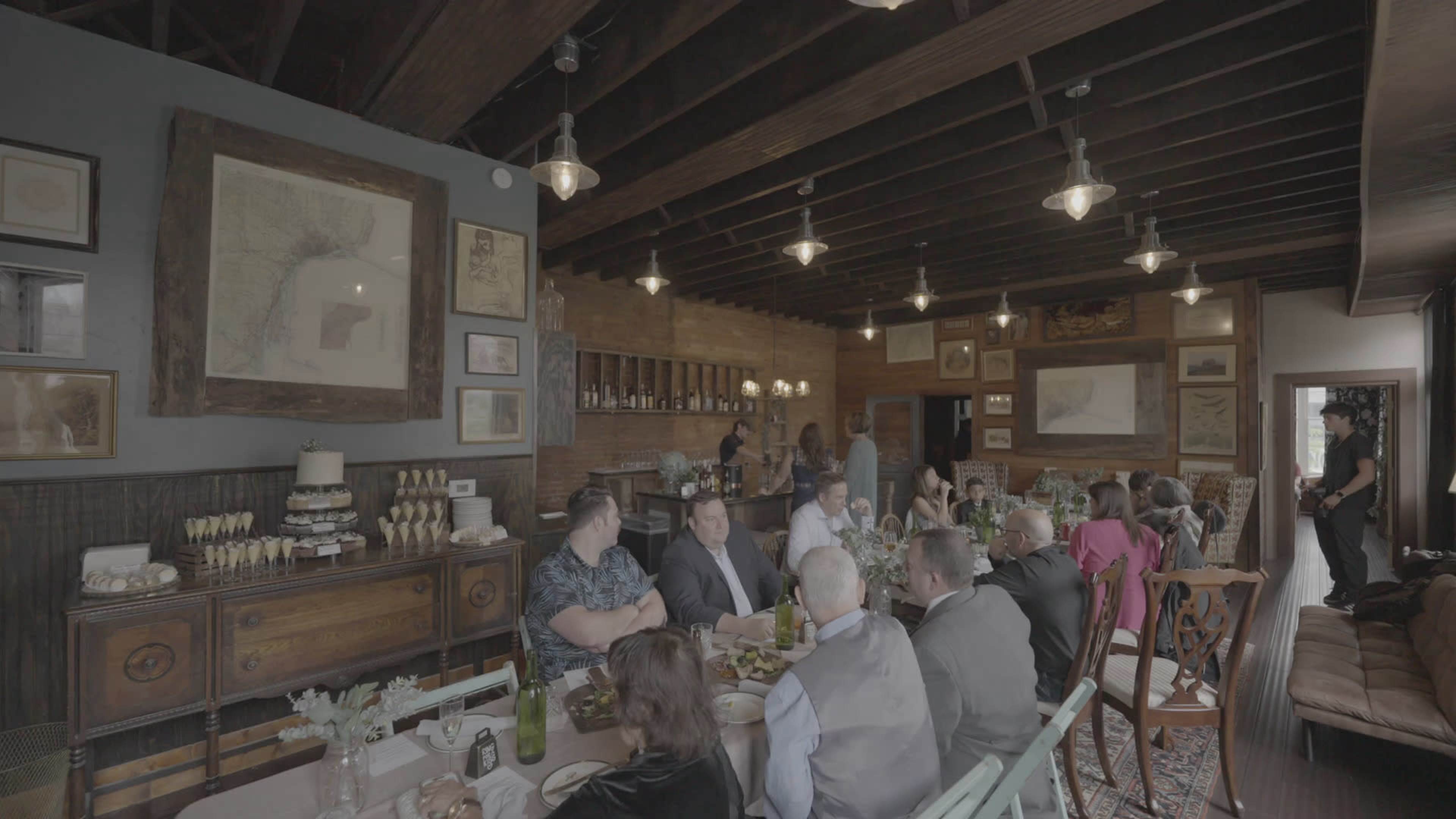 A group of people is gathered around a long table in a rustic dining area decorated with framed maps and vintage artwork.