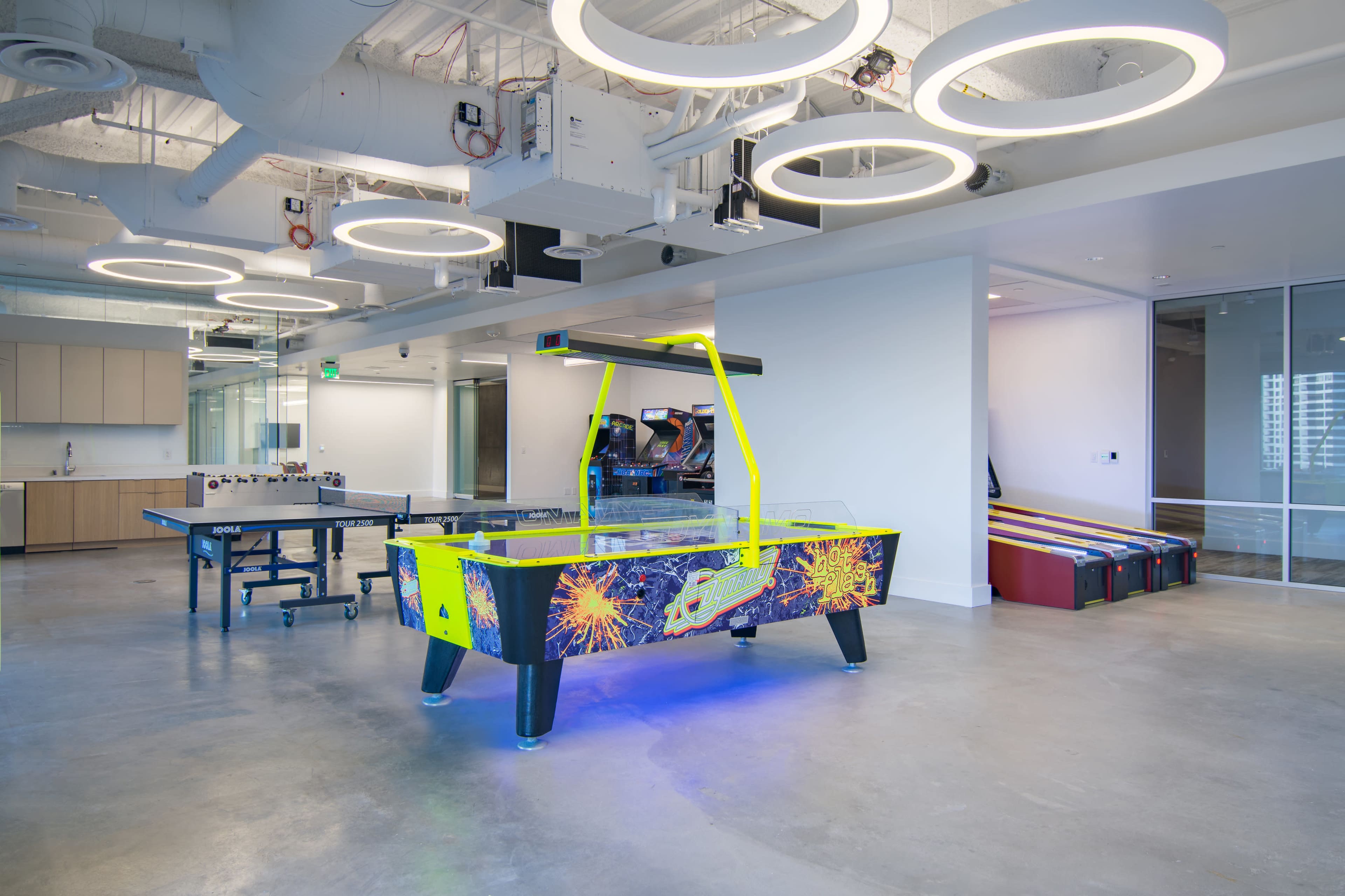 A modern office space featuring a brightly colored air hockey table, a ping pong table, and multiple arcade machines in a well-lit environment.