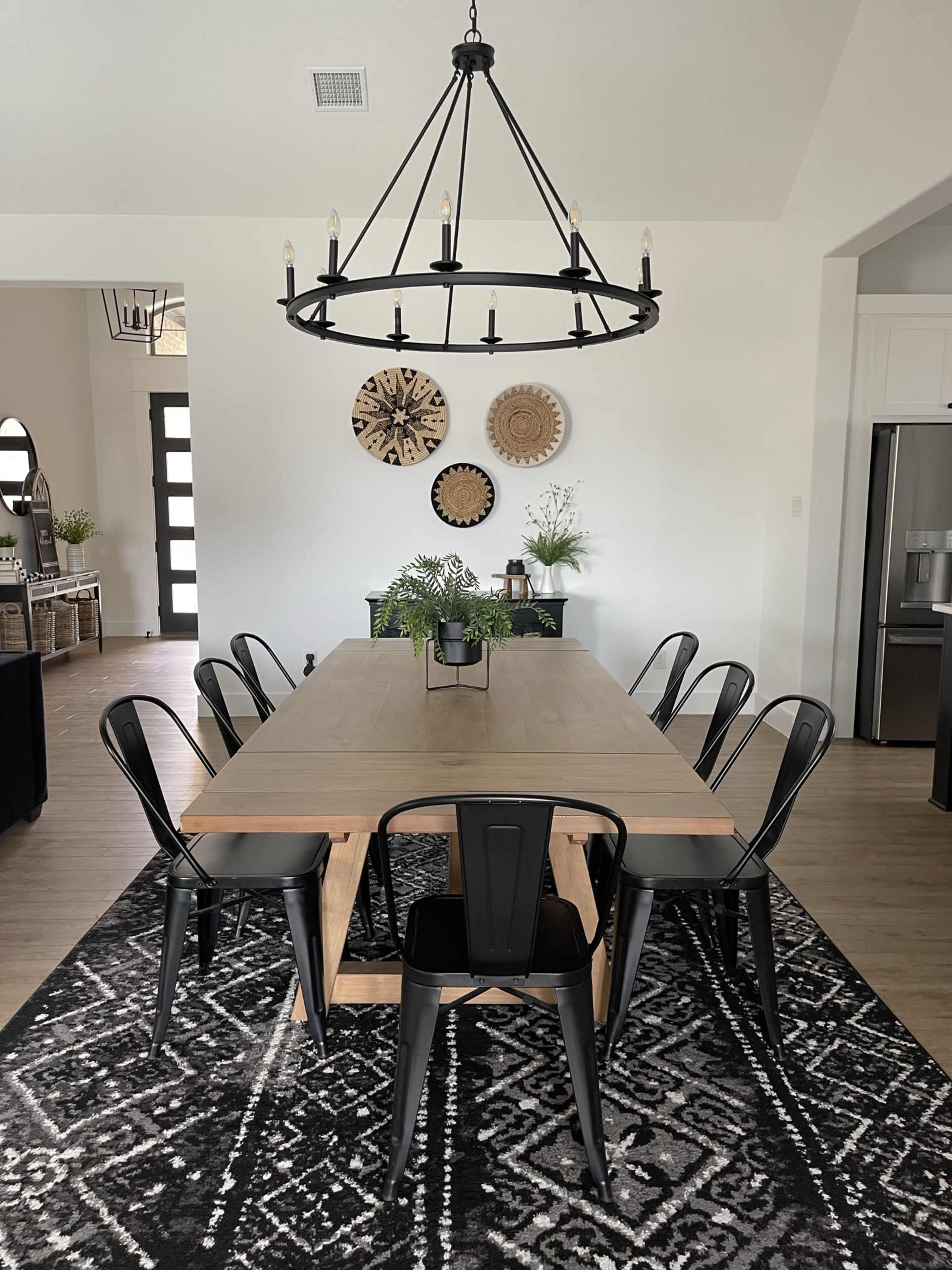 A spacious dining room features a large wooden table surrounded by black chairs, with a chandelier above and decorative wall plates in the background.