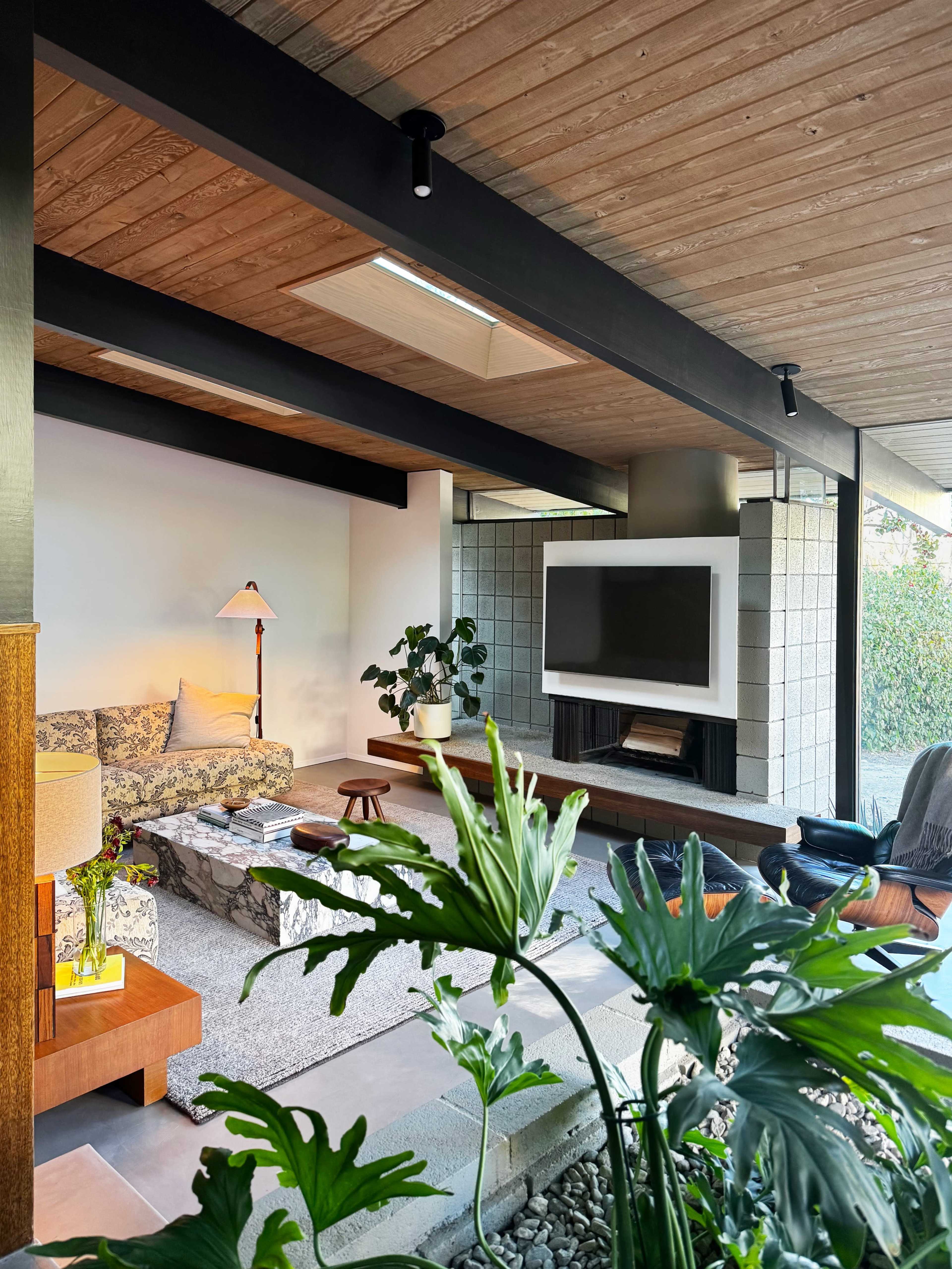 The image shows a modern living room featuring a sofa, a coffee table, a television mounted on a wall, and indoor plants decorating the space.
