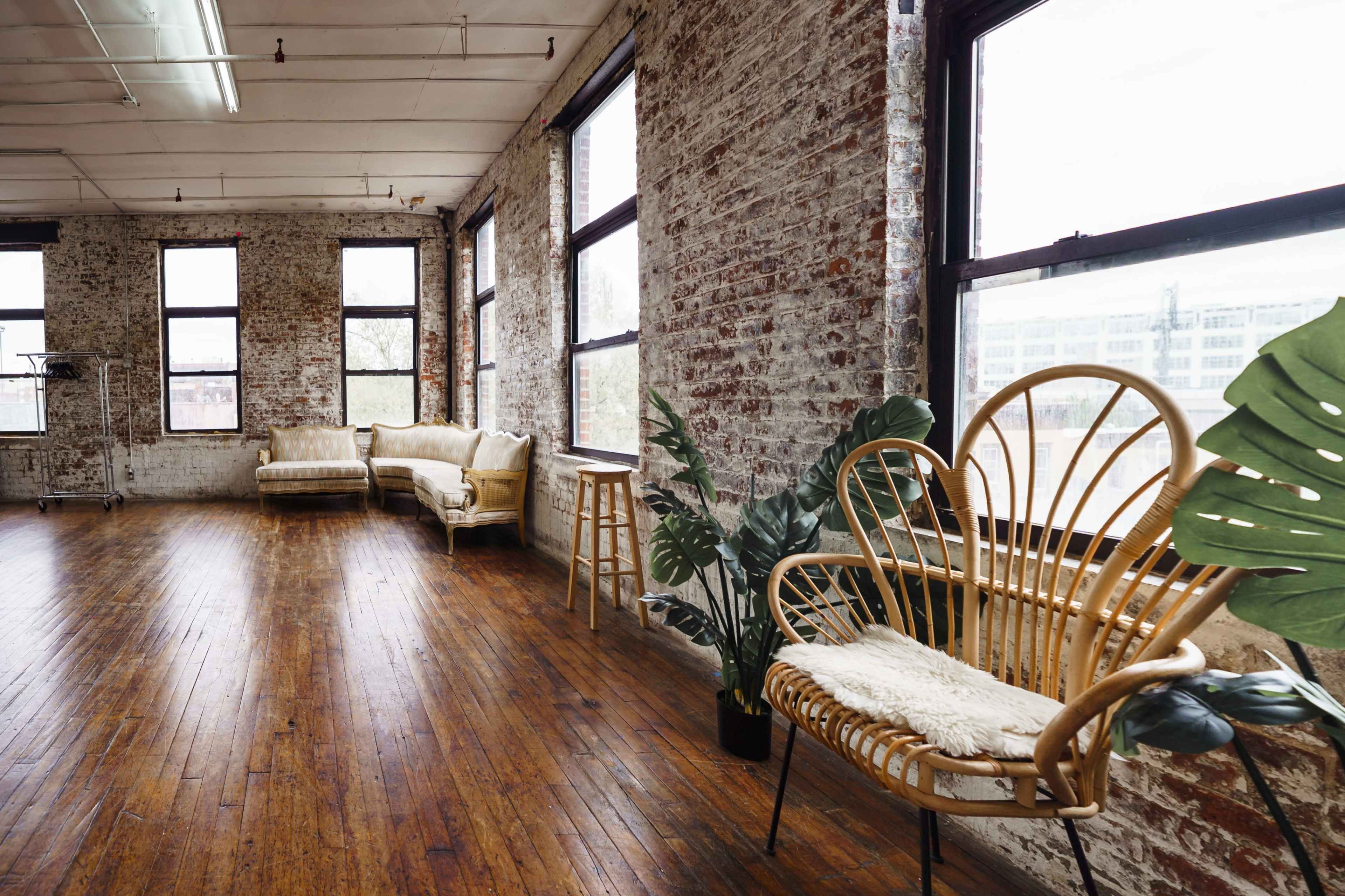 The scene depicts an airy interior space with exposed brick walls, large windows, wooden flooring, and various seating arrangements, including a rattan chair and a beige couch.