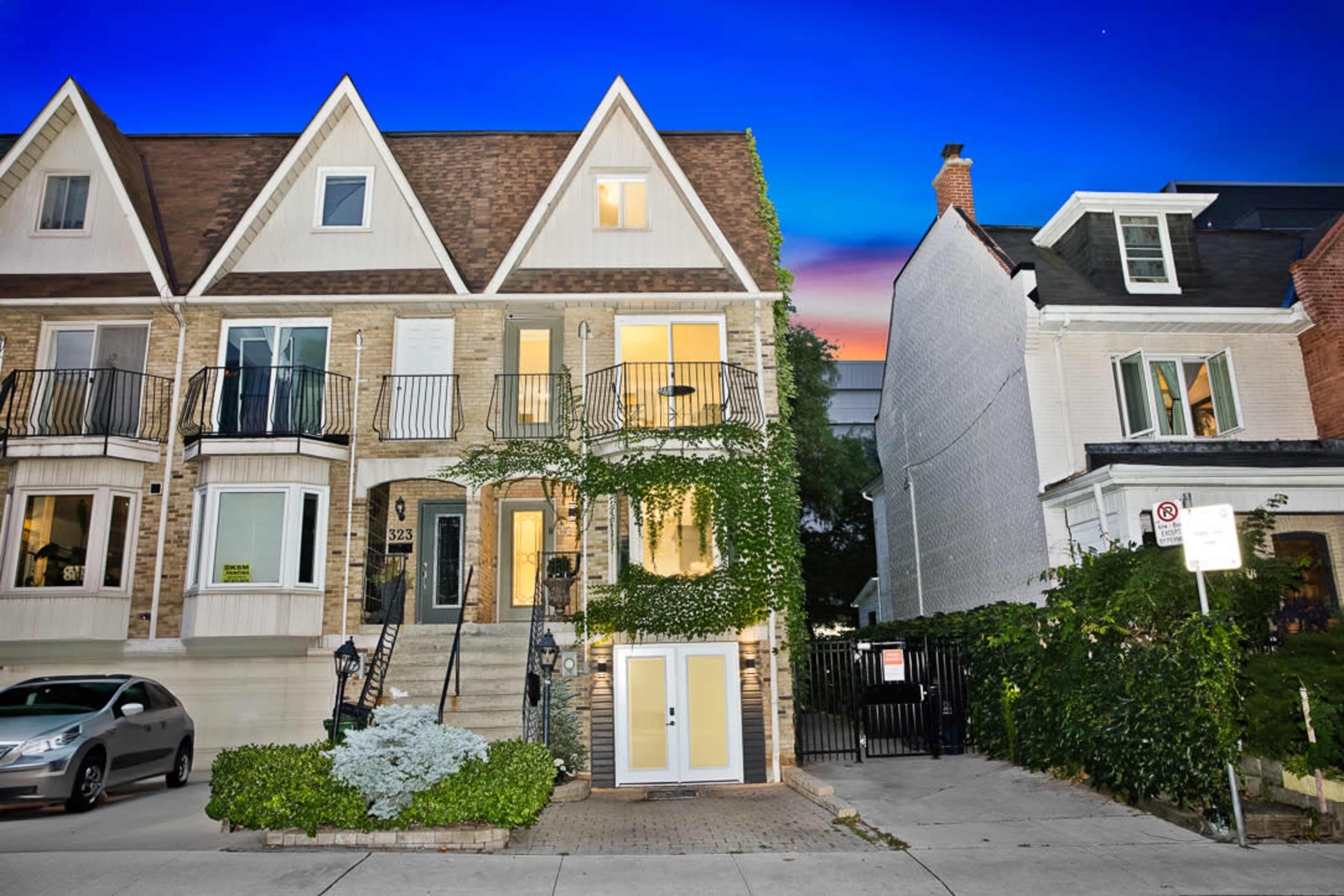 The image shows a multi-story brick townhouse with a triangular roof and balconies, surrounded by greenery and a clear blue sky at dusk.