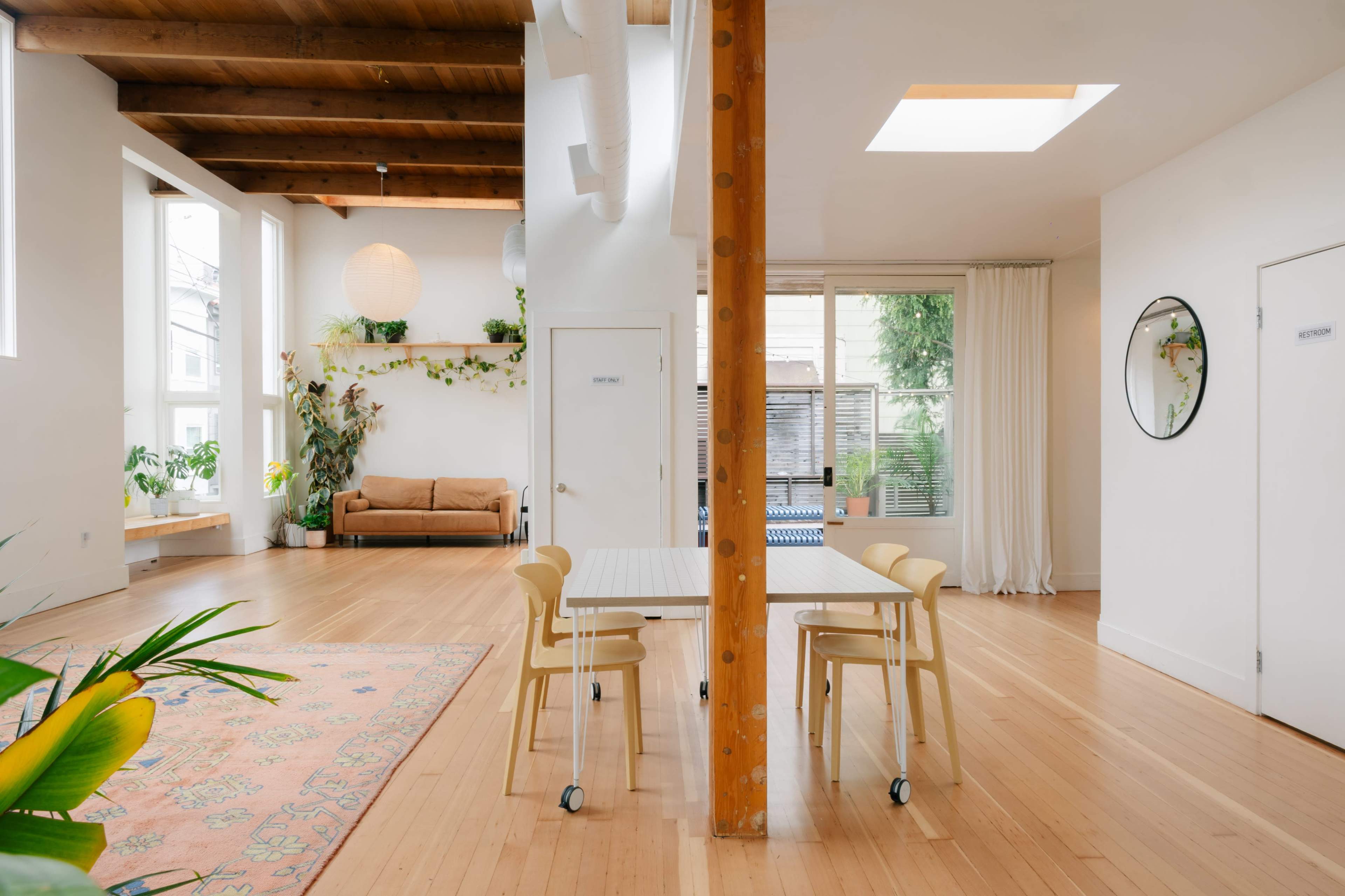 The image shows a bright, spacious room with wooden beams, a table and chairs, a couch, and several plants arranged throughout.