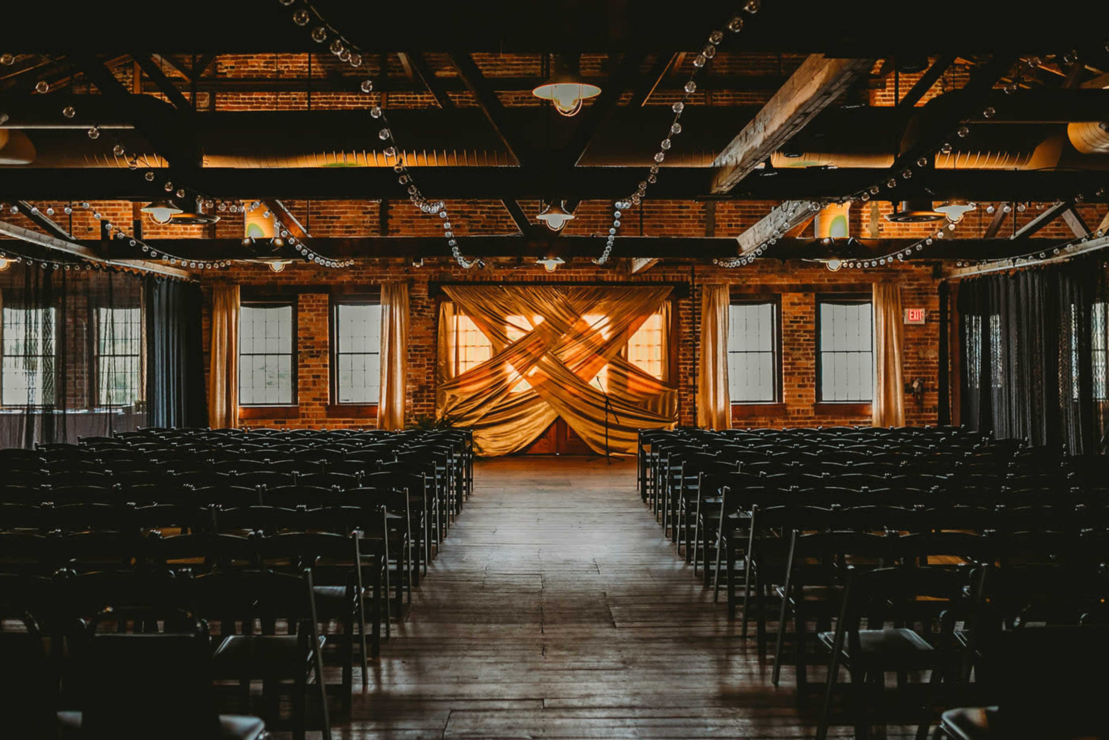 An indoor event space with rows of black chairs facing a draped stage adorned with golden fabric and decorative lighting.