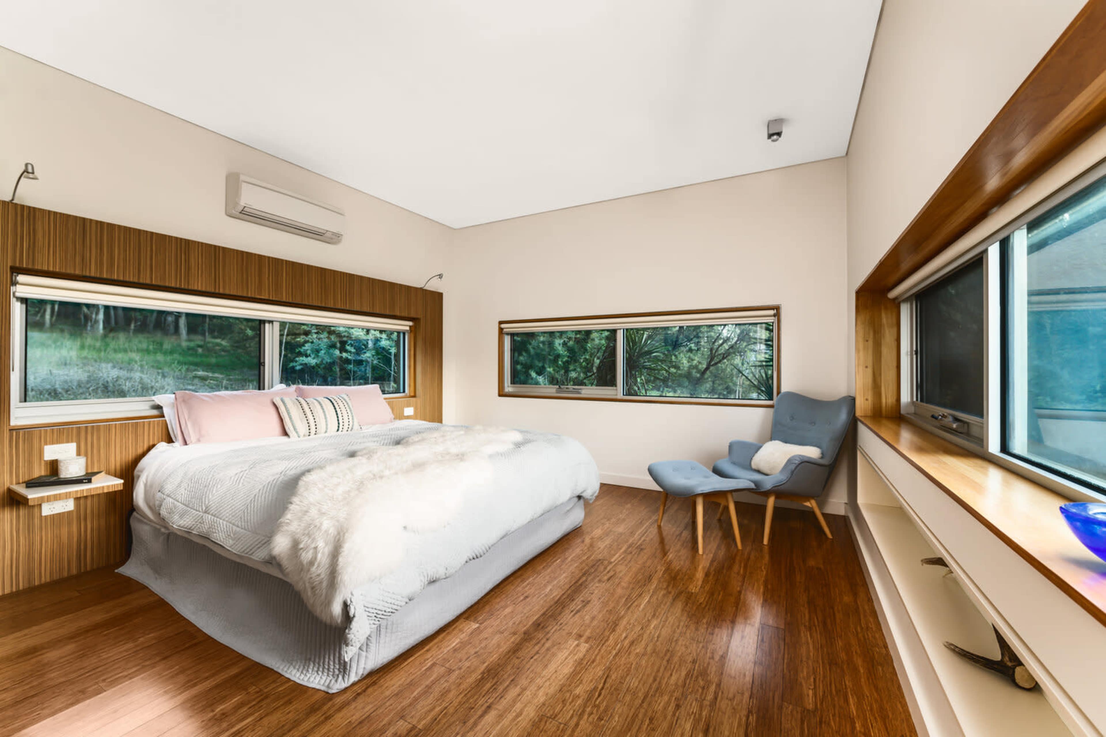 A modern bedroom with a large bed, wooden accents, and large windows overlooking a wooded area.