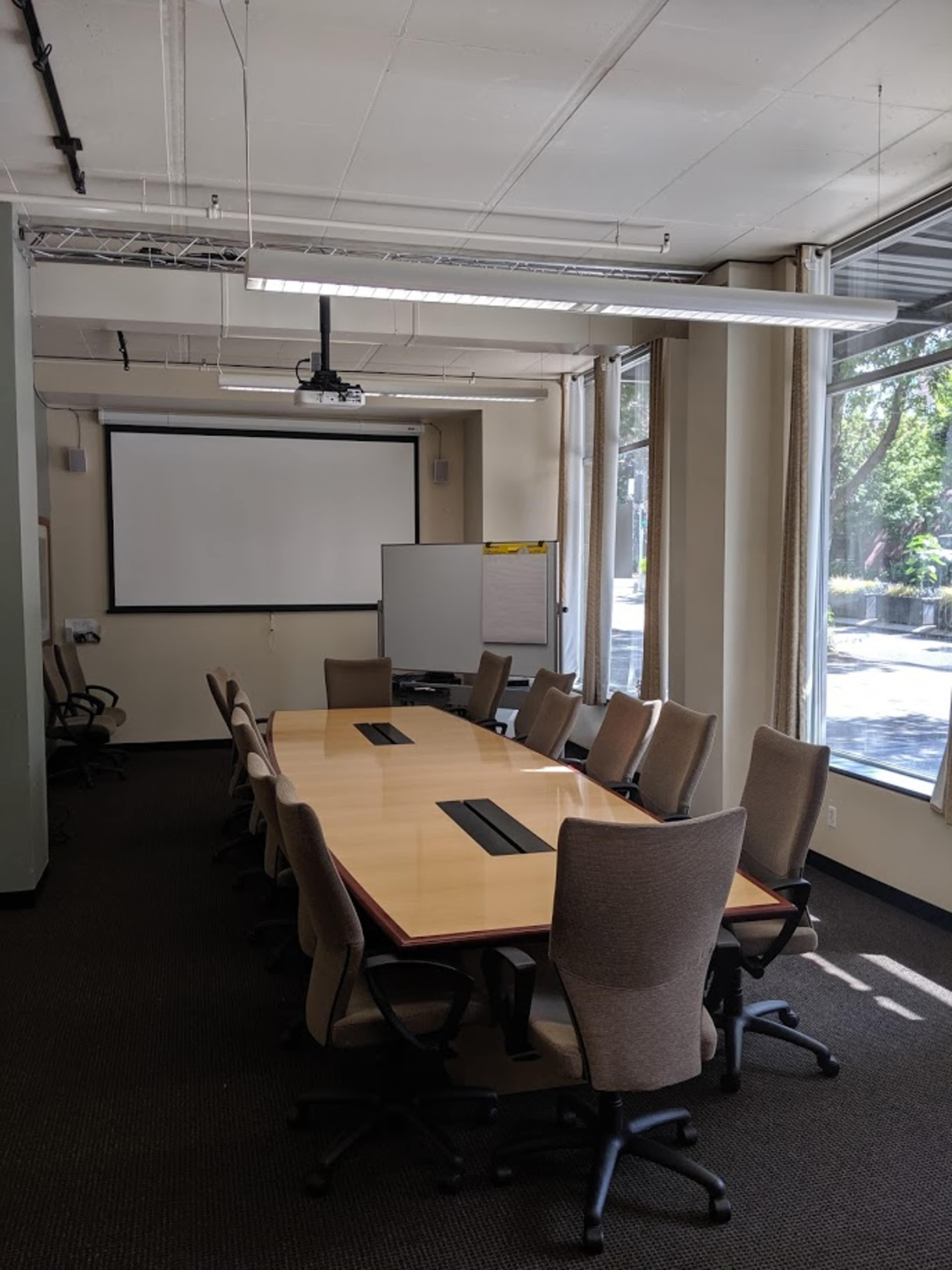 A large conference room features a long table with several chairs arranged around it, a projector screen on the front wall, and windows providing natural light.