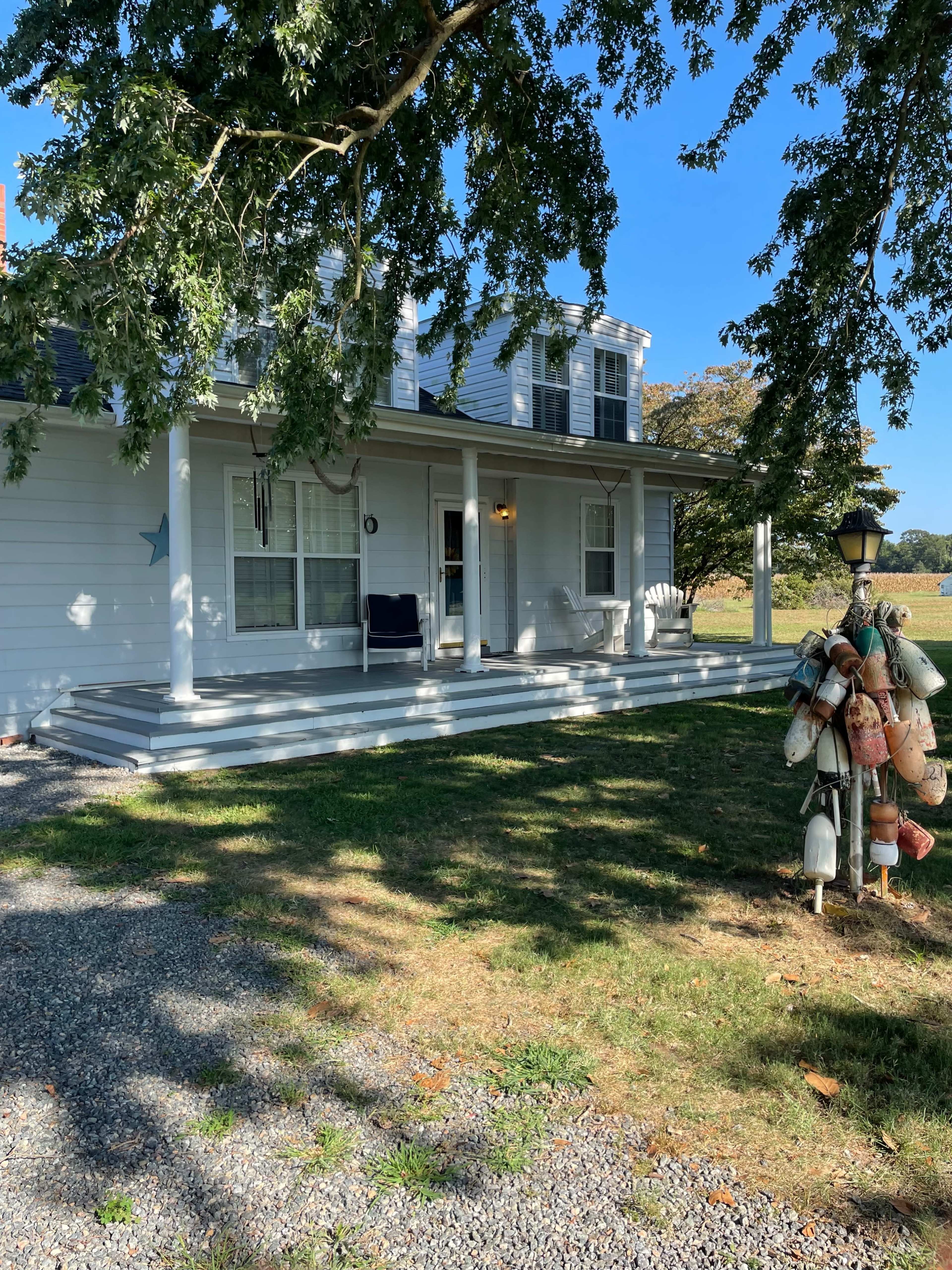 A two-story white house with a porch, surrounded by grass and trees, along with a display of buoys on the right.