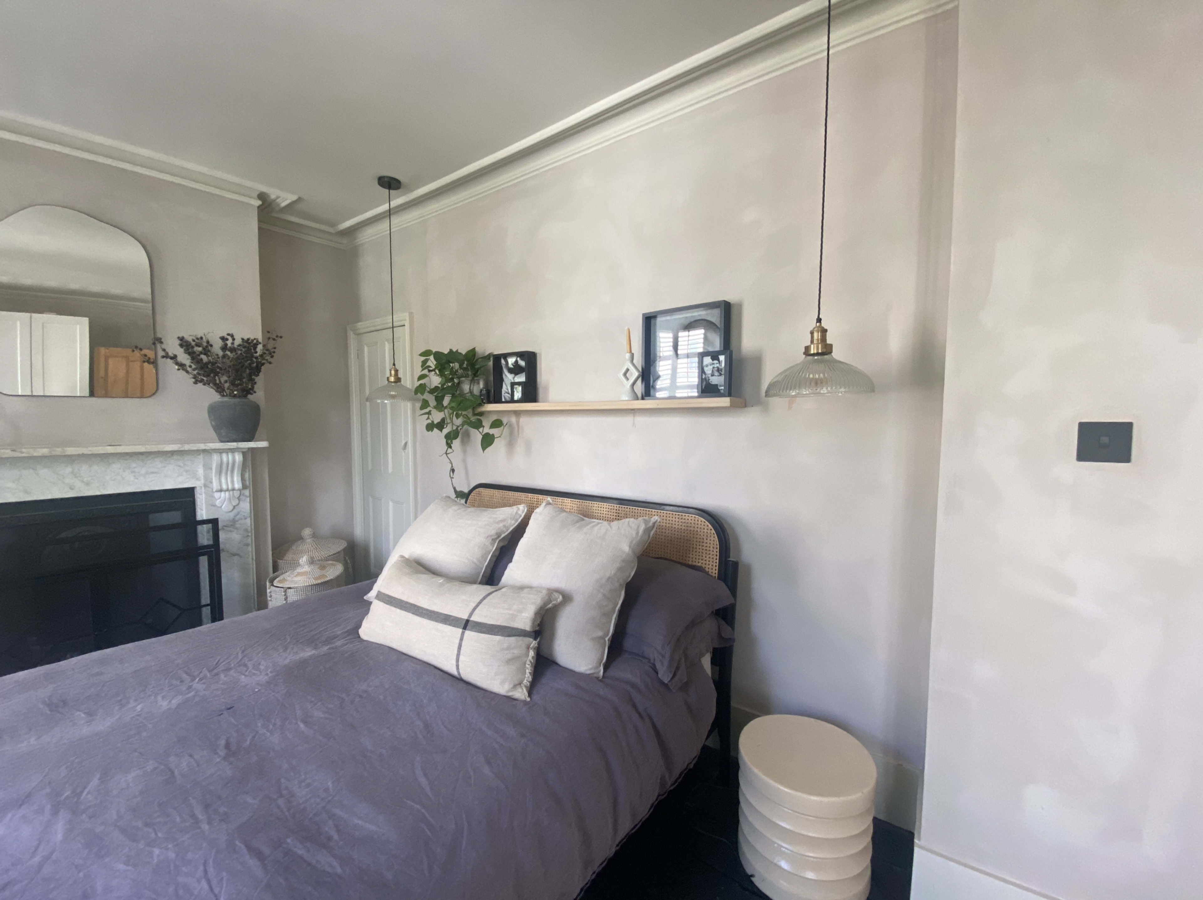 A bedroom featuring a bed with dark bedding, a bedside table, a wall shelf with decorative items, and a fireplace.