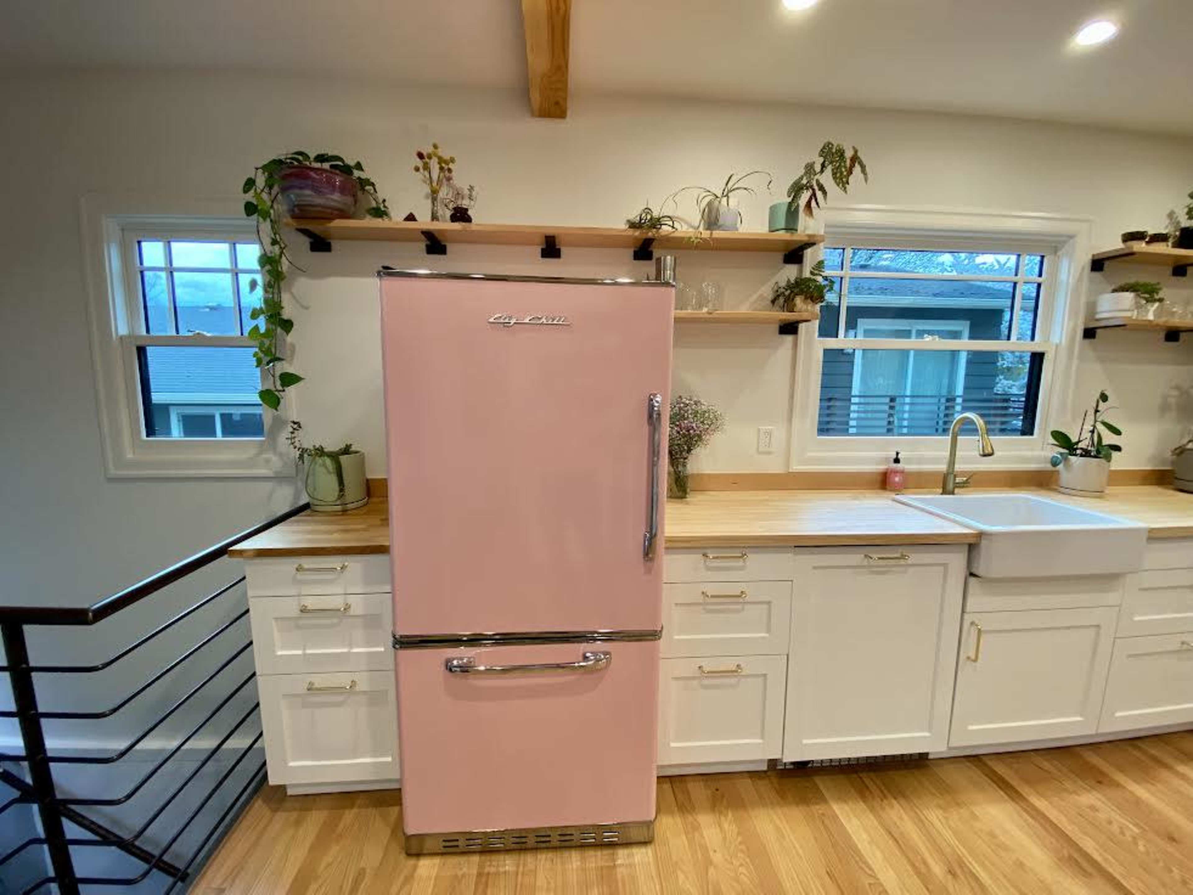 A pink retro-style refrigerator stands against a wooden countertop in a modern kitchen, surrounded by white cabinets and various plants on shelves.