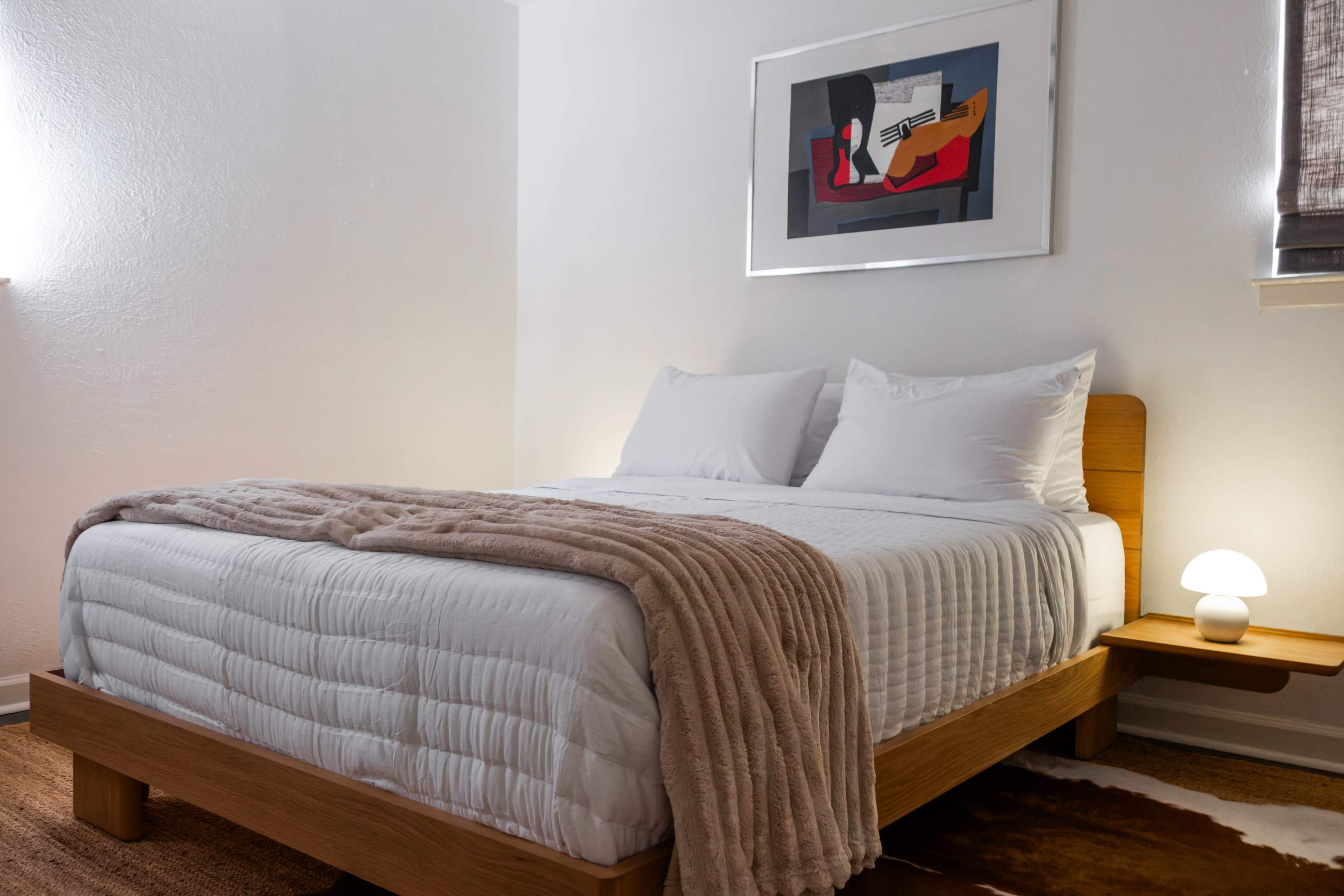 The image shows a neatly arranged bedroom with a wooden bed frame, a white quilted comforter, and artwork hanging on the wall above the bed.