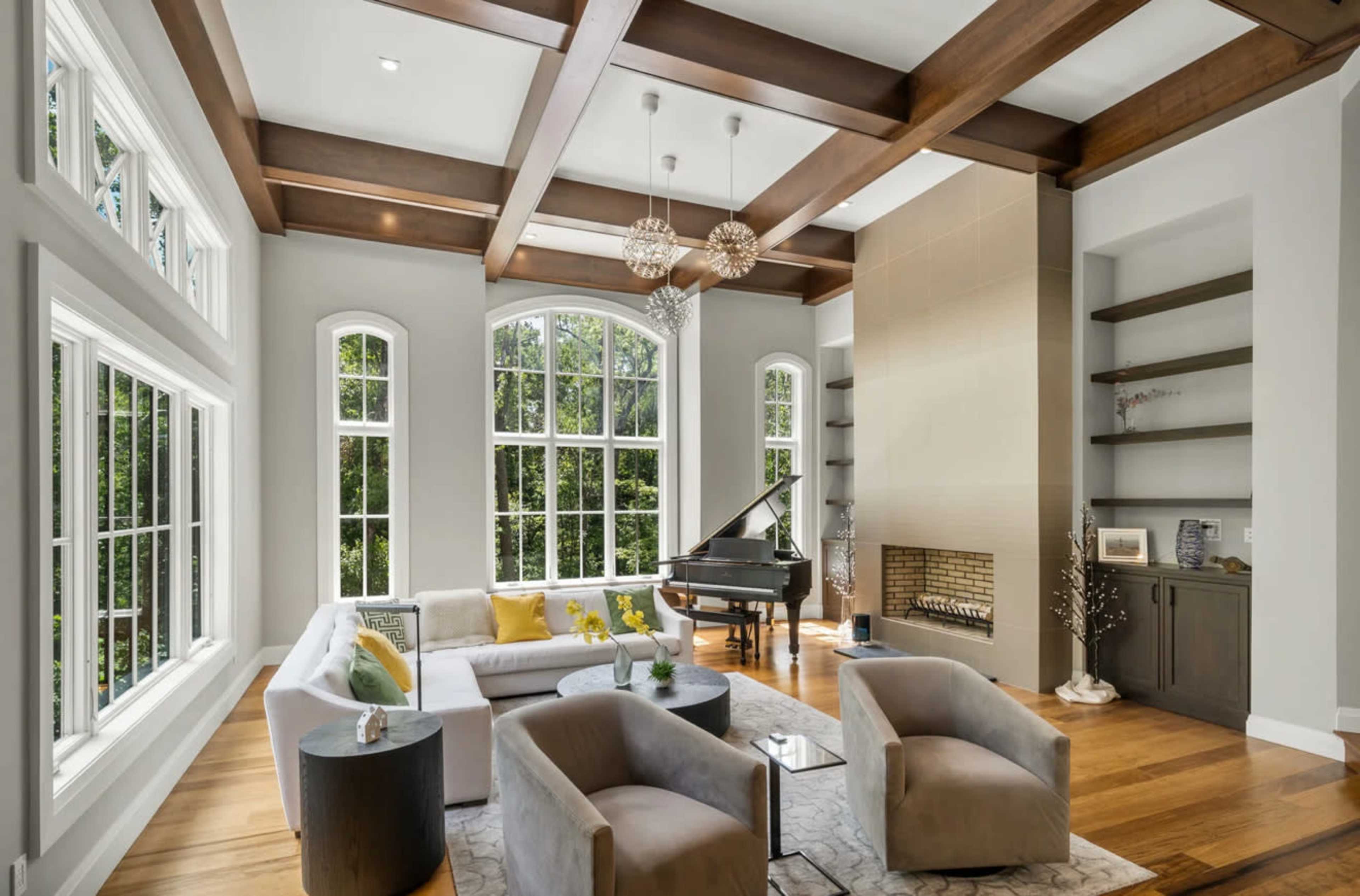 A spacious living room features large windows, wooden beams, a grand piano, and modern furniture arranged around a fireplace.