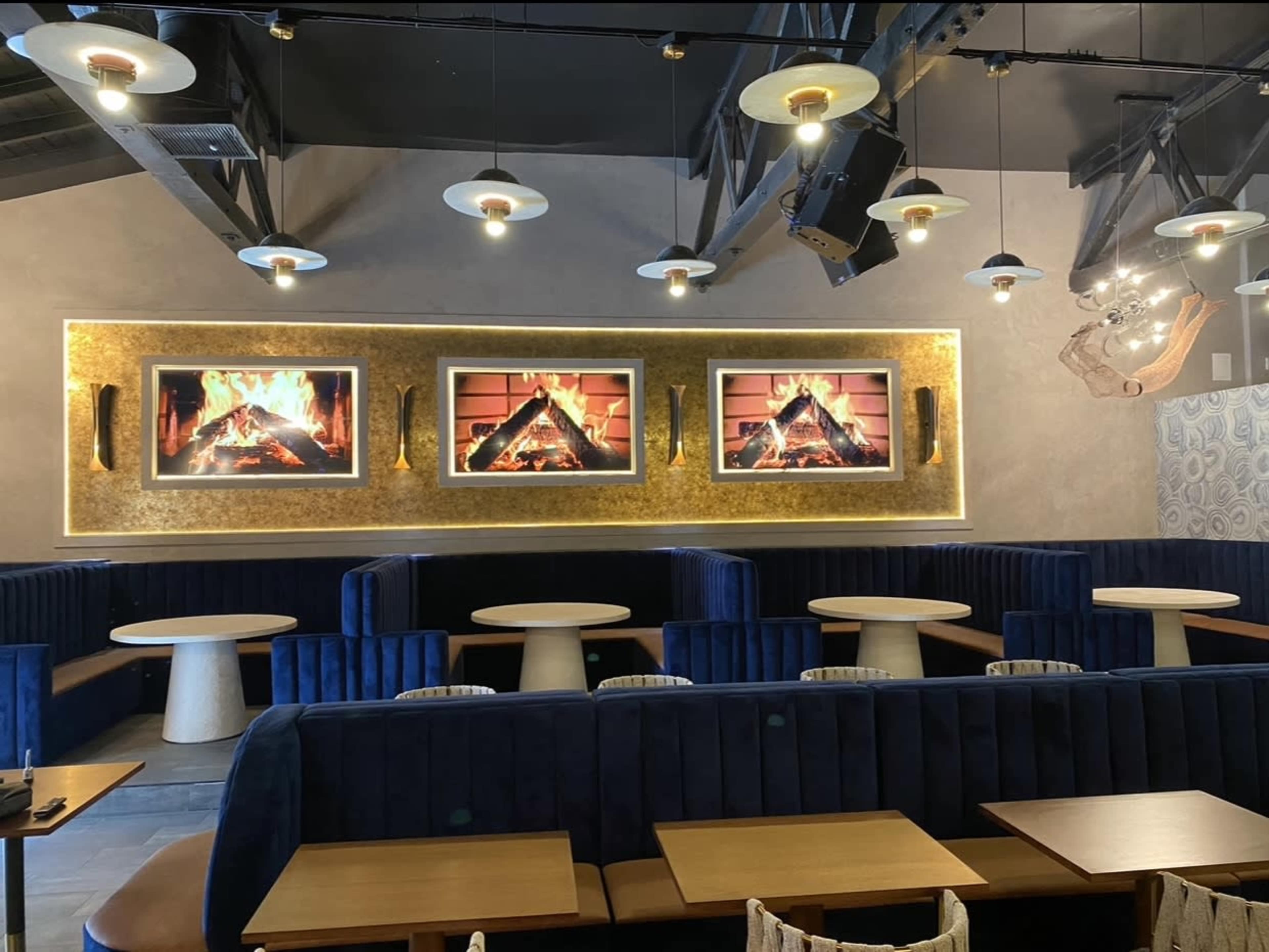 A modern restaurant interior with blue booths, white tables, and three large frames displaying simulated fireplace visuals on the wall.