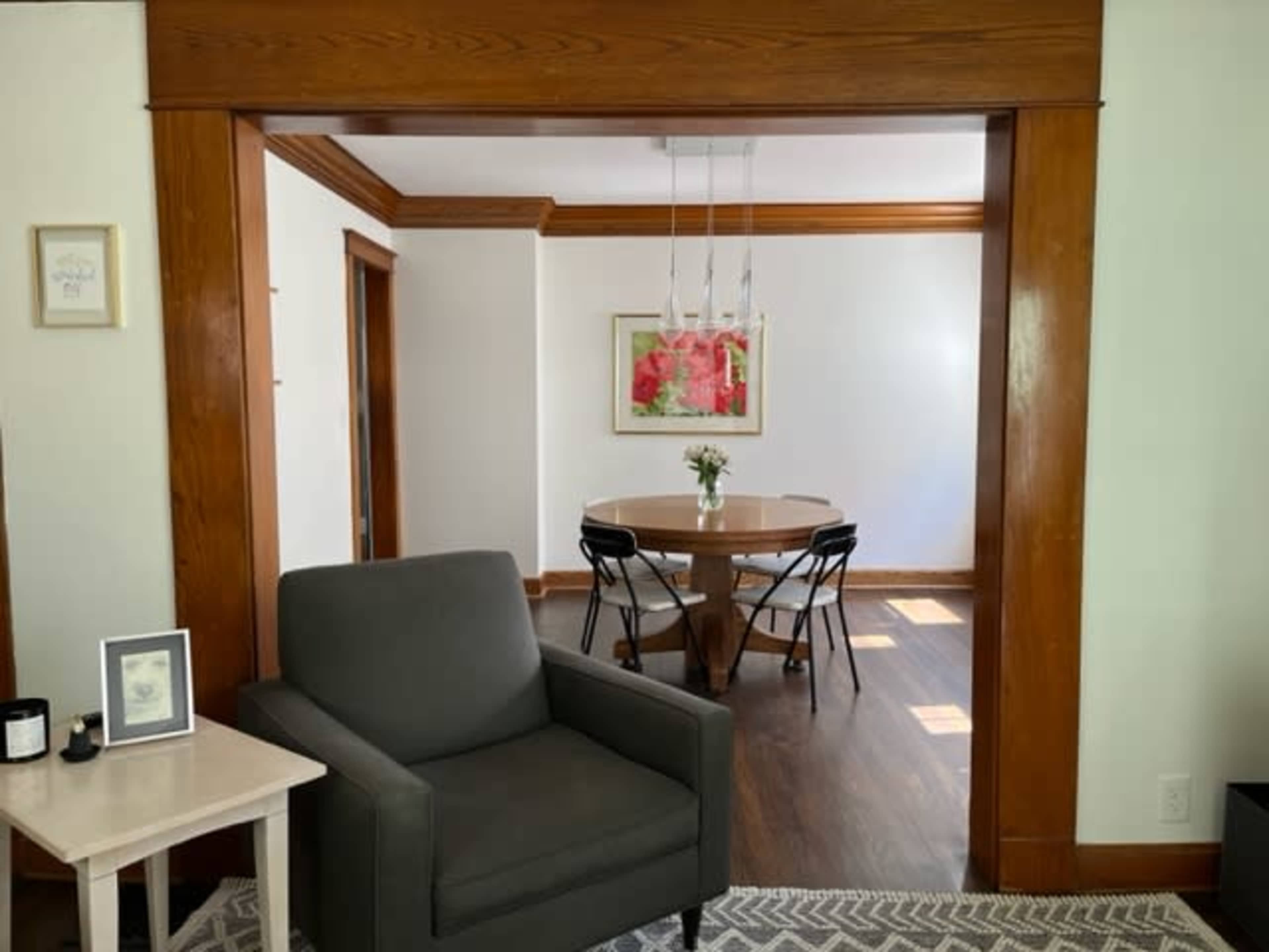 The image shows a living room area with a gray armchair and a small table, leading to a dining area featuring a round table and a floral painting on the wall.